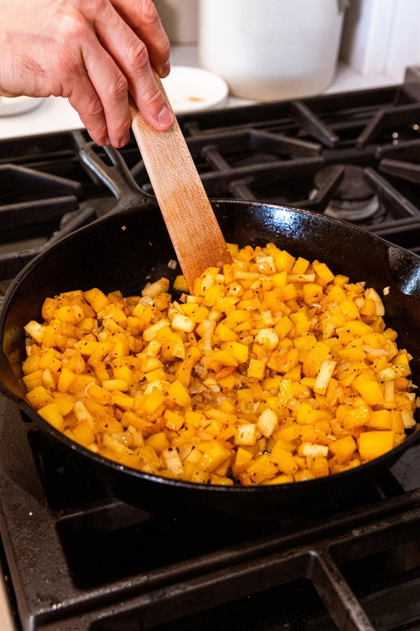 Mixture of squash and apples and onion cooking together in cast iron skillet on stovetop with wooden spatula mixing everything together.