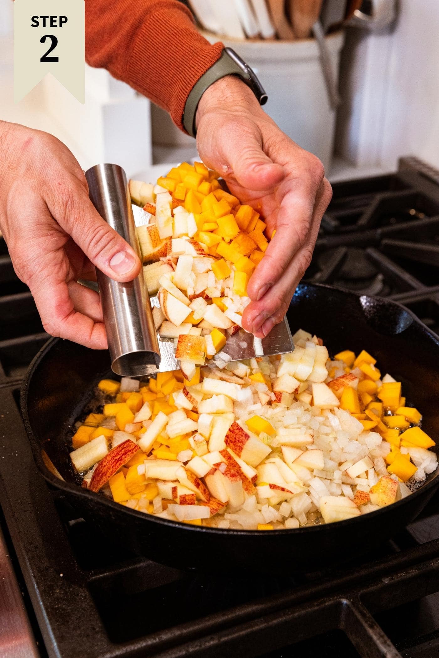 Hand holding chunks of butternut squash and apples pouring into a cast iron skillet on stovetop.