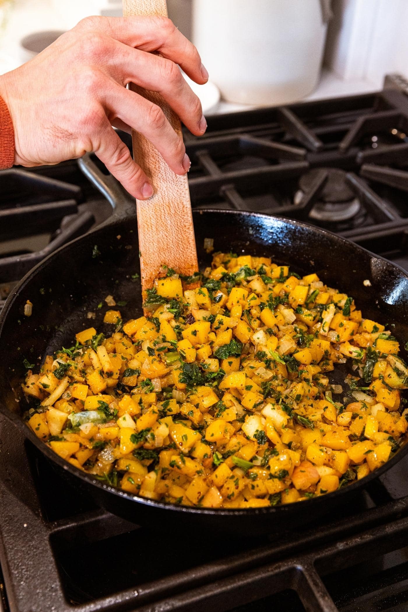 Hand using wooden spatula to mix together pieces of butternut squash and kale in black cast iron skillet on stovetop.