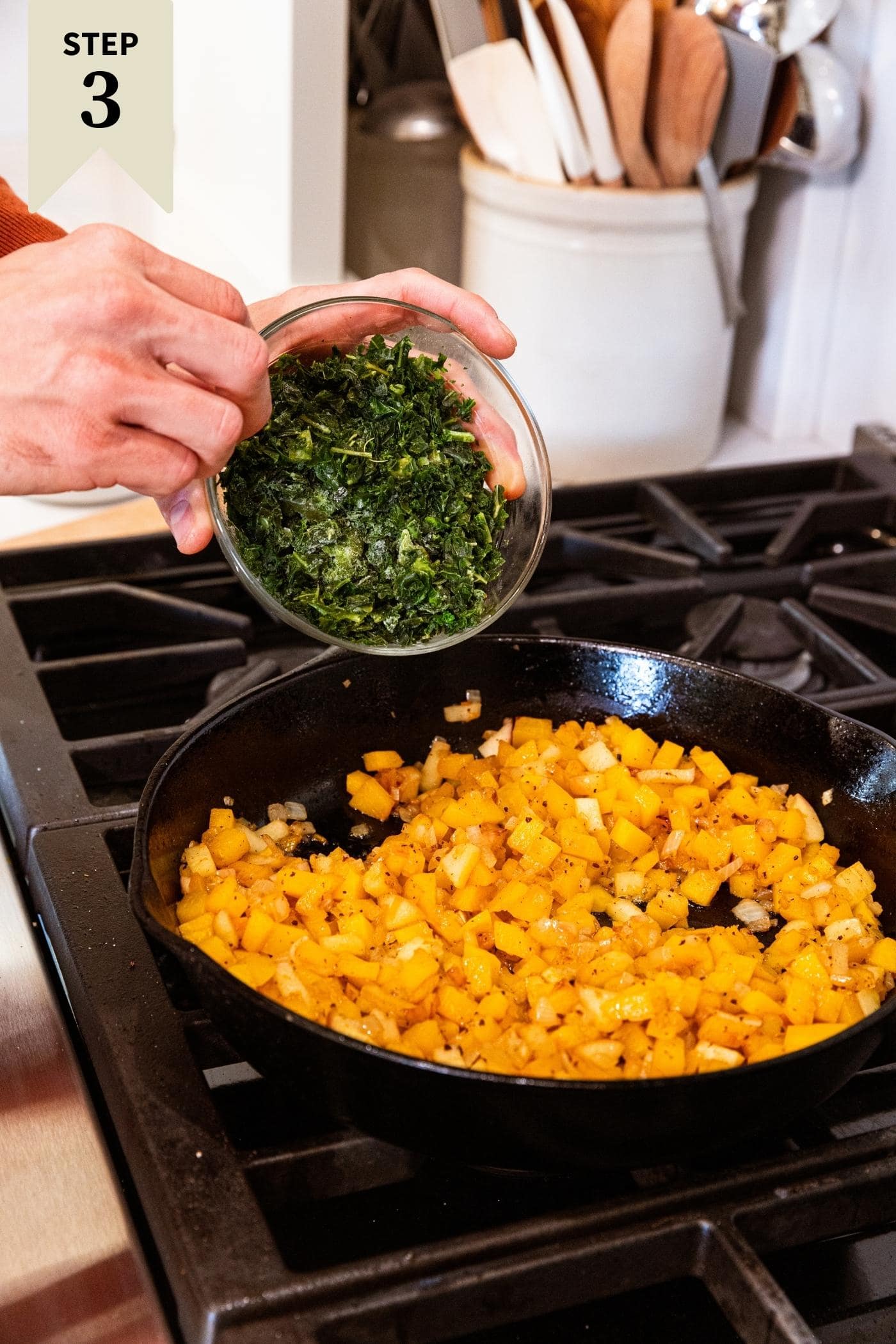 Hand holding small glass bowl filled with frozen kale about to pour it into a cast iron skillet on stovetop filled with cooked butternut squash pieces.