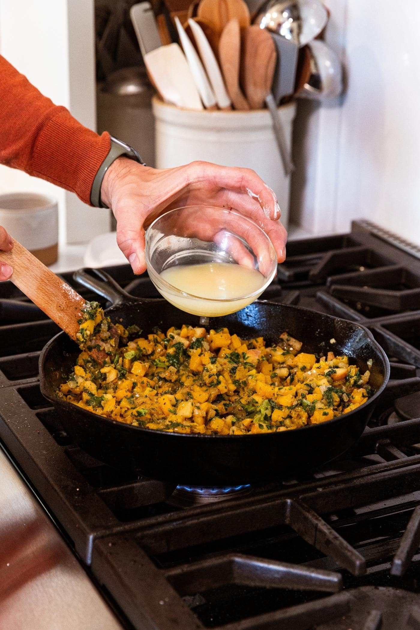 Hand holding glass bowl filled with light yellow colored chicken stock about to pour into cast iron skillet filled with diced butternut squash and kale.