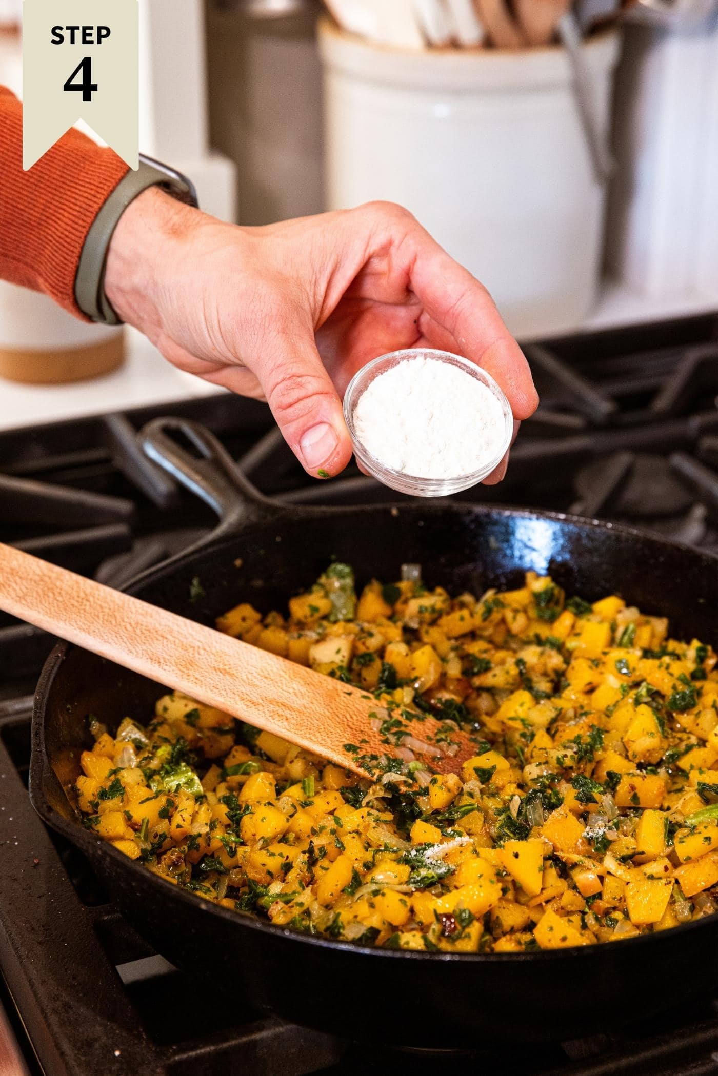 Hand holding small glass bowl filled with flour about to pour into a black cast iron skillet filled with butternut squash and kale mixture on the stove after cooking for some time.
