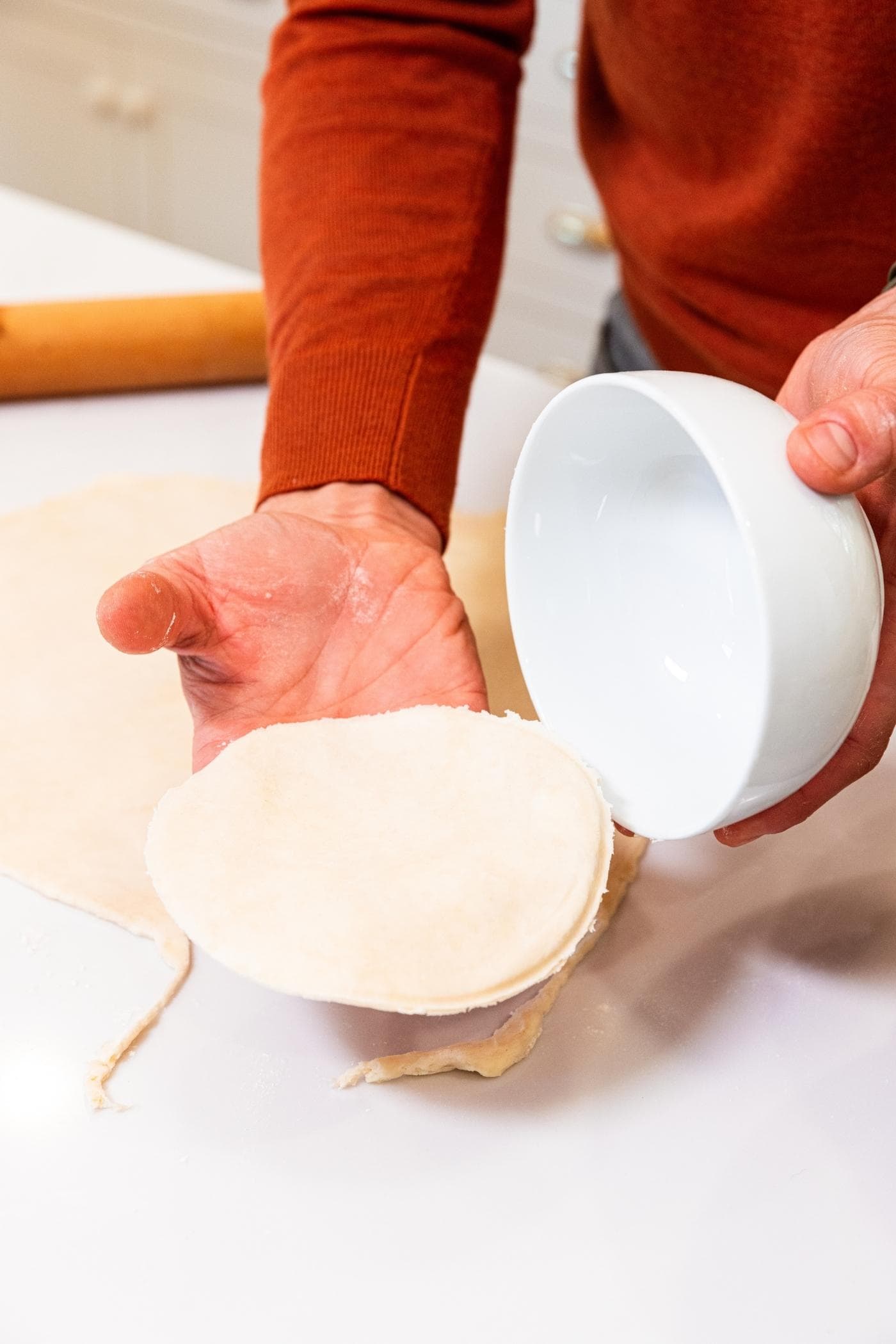 Hand pulling white bowl off of round of dough after being sliced from rest of dough with white countertop underneath.