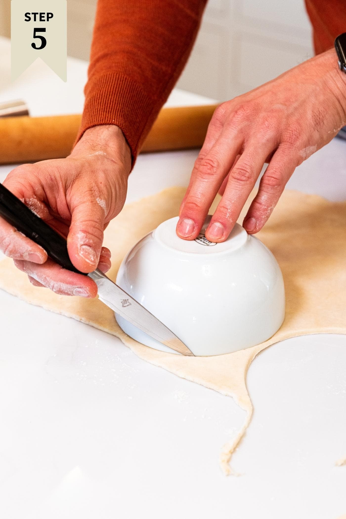 Hand using knife to cut out circle from dough using a white bowl laid upside down on top of dough all on white countertop.