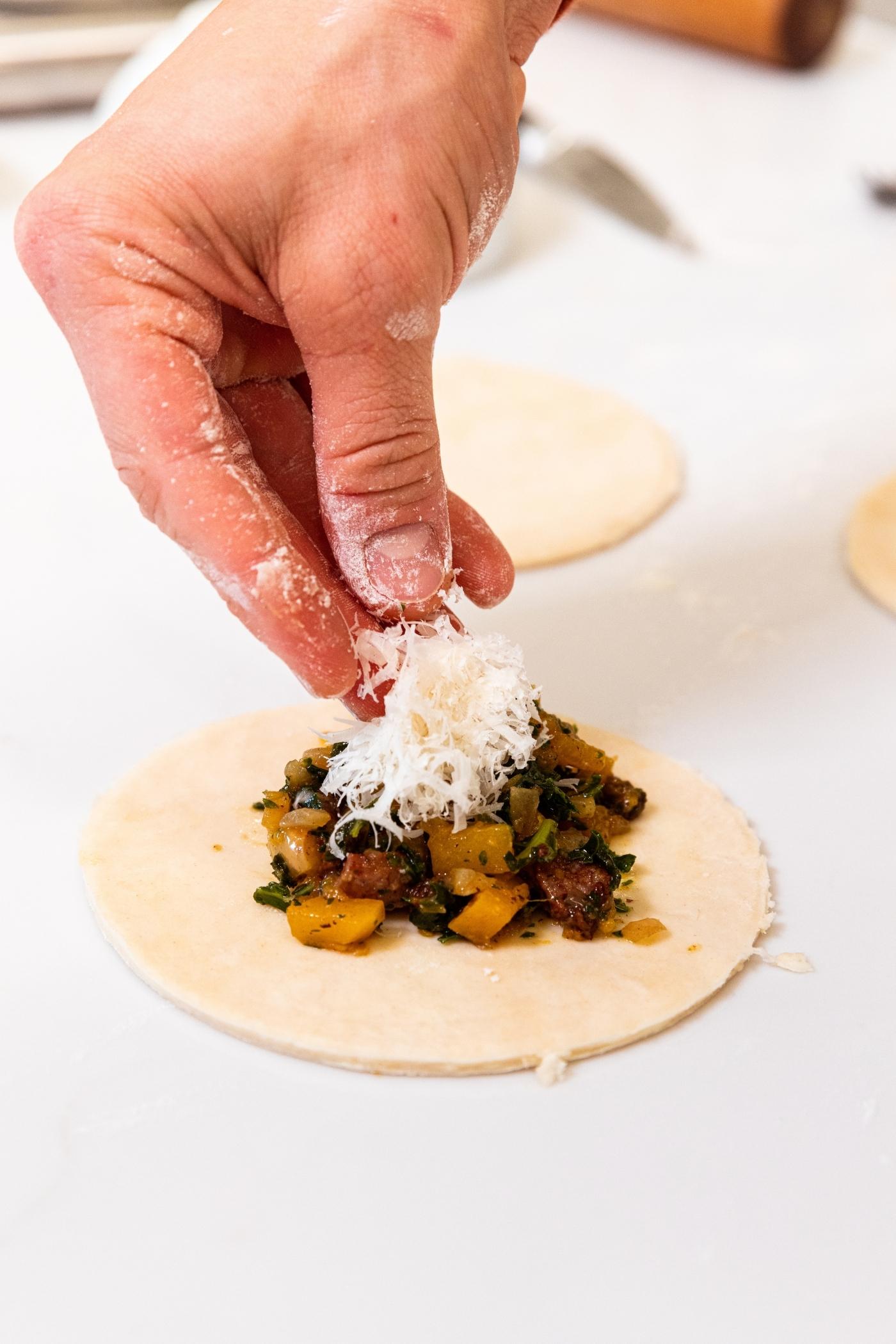 Hand dropping a sprinkling of Parmesan cheese on top of butternut squash mixture on top of round of pie dough on white countertop.