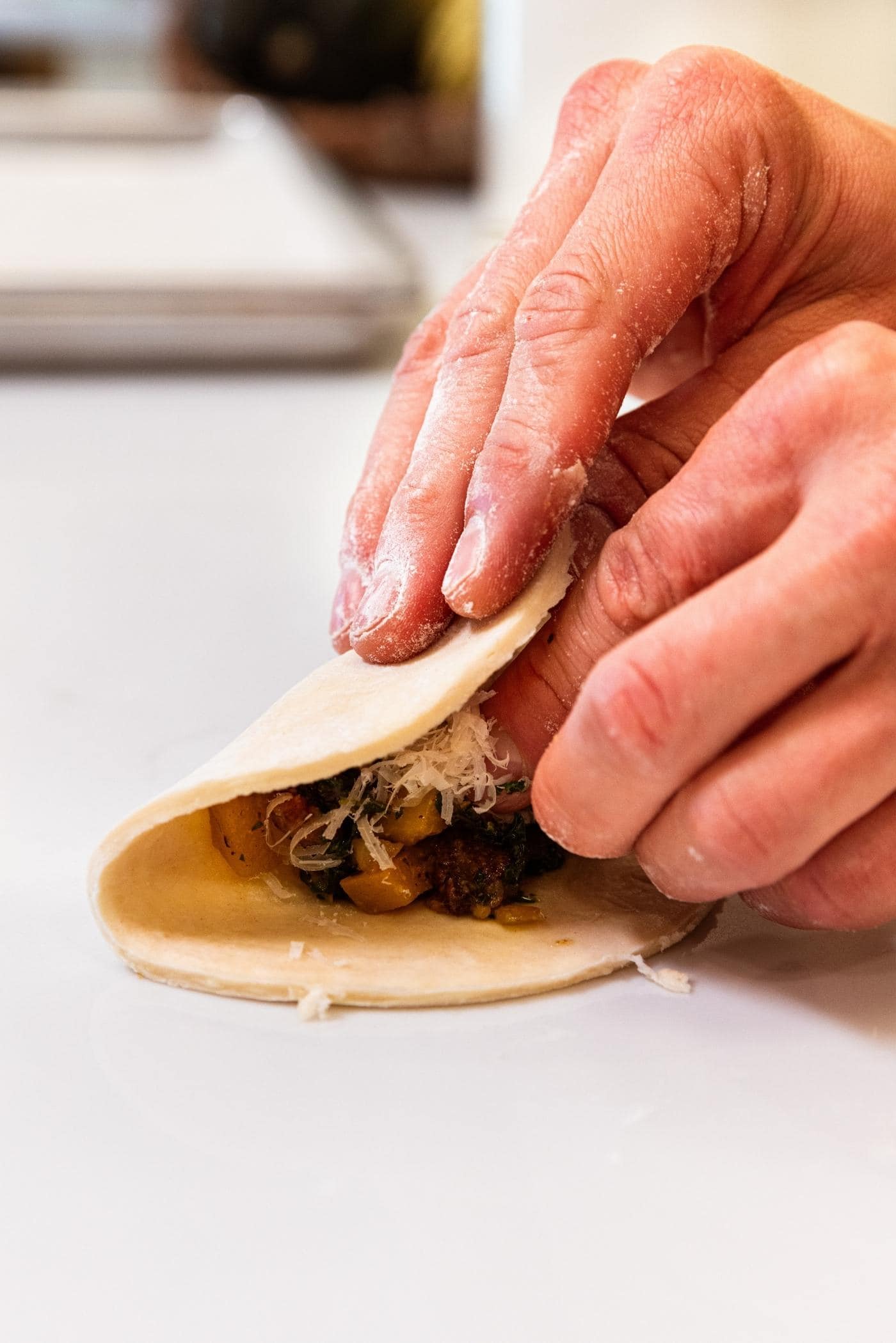 Two hands folding over half of a dough circle on top of a dollop of butternut squash and sausage mixture all sitting on white countertop.