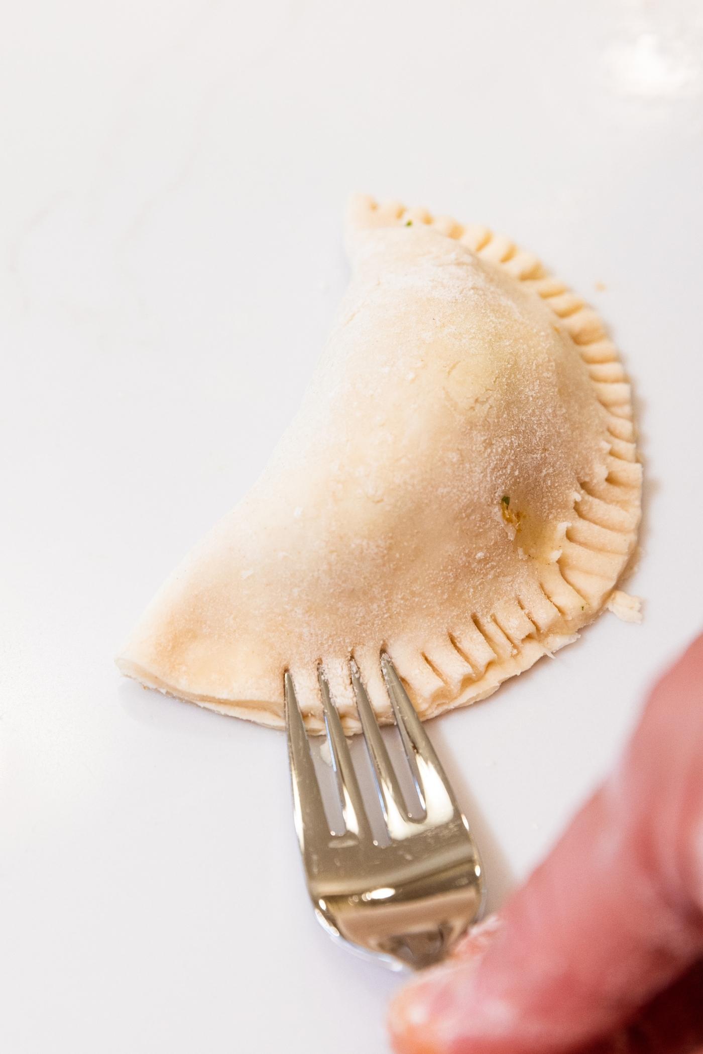 Fork pressing tines down into dough of half moon shaped hand pie sitting on white countertop.