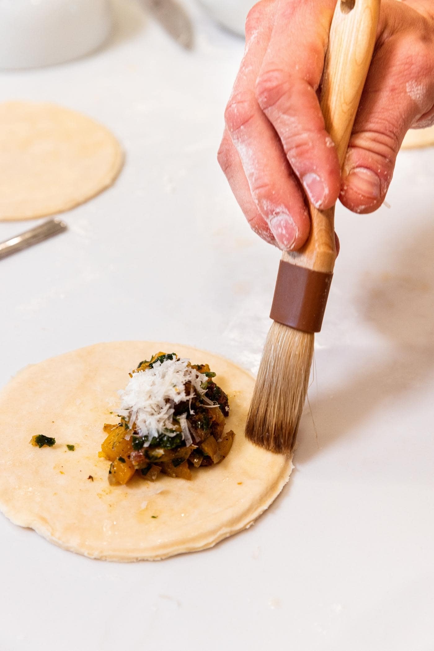 Hand using brush to use an egg wash to brush on one side of a circle of dough sitting on white countertop with small dollop of butternut squash and sausage mixture with Parmesan cheese on top.
