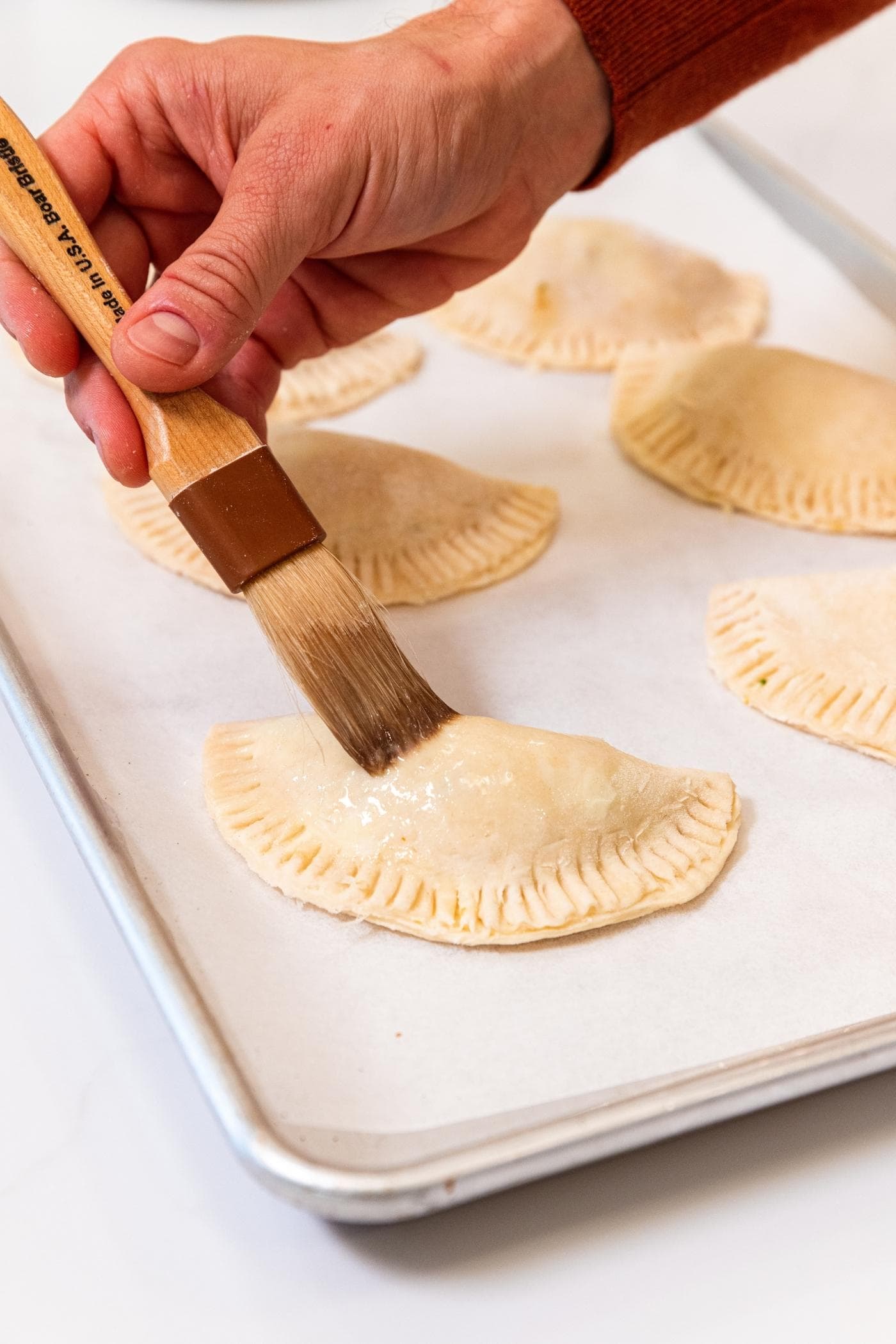 Hand using a brush to put an egg wash on the top of a half moon shaped hand pie sitting on a parchment-lined baking sheet with more hand pies in background.