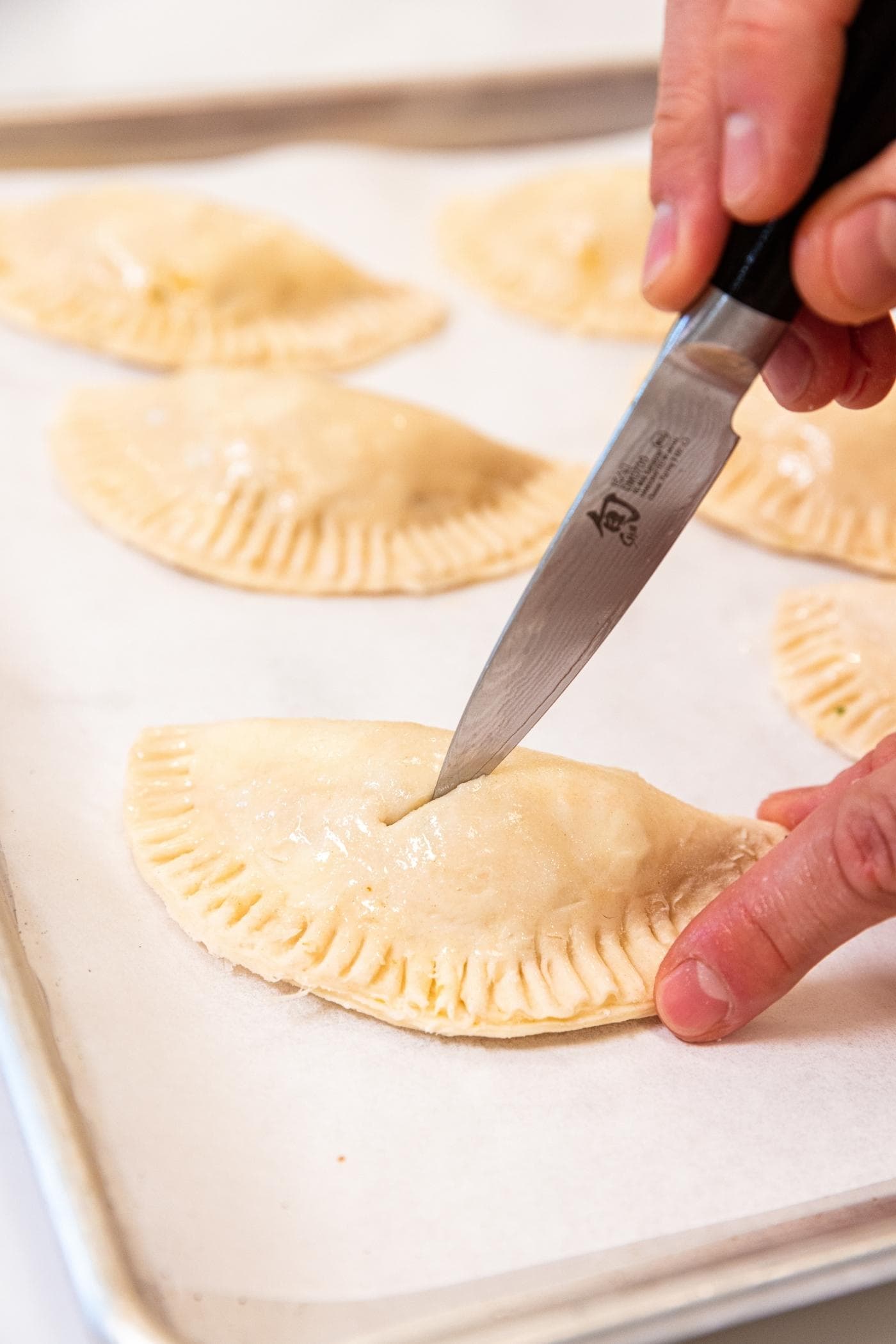 Hand using a knife to slice a vent hole in the top of a half shaped hand pie sitting on a parchment-lined baking sheet with more hand pies in the background.