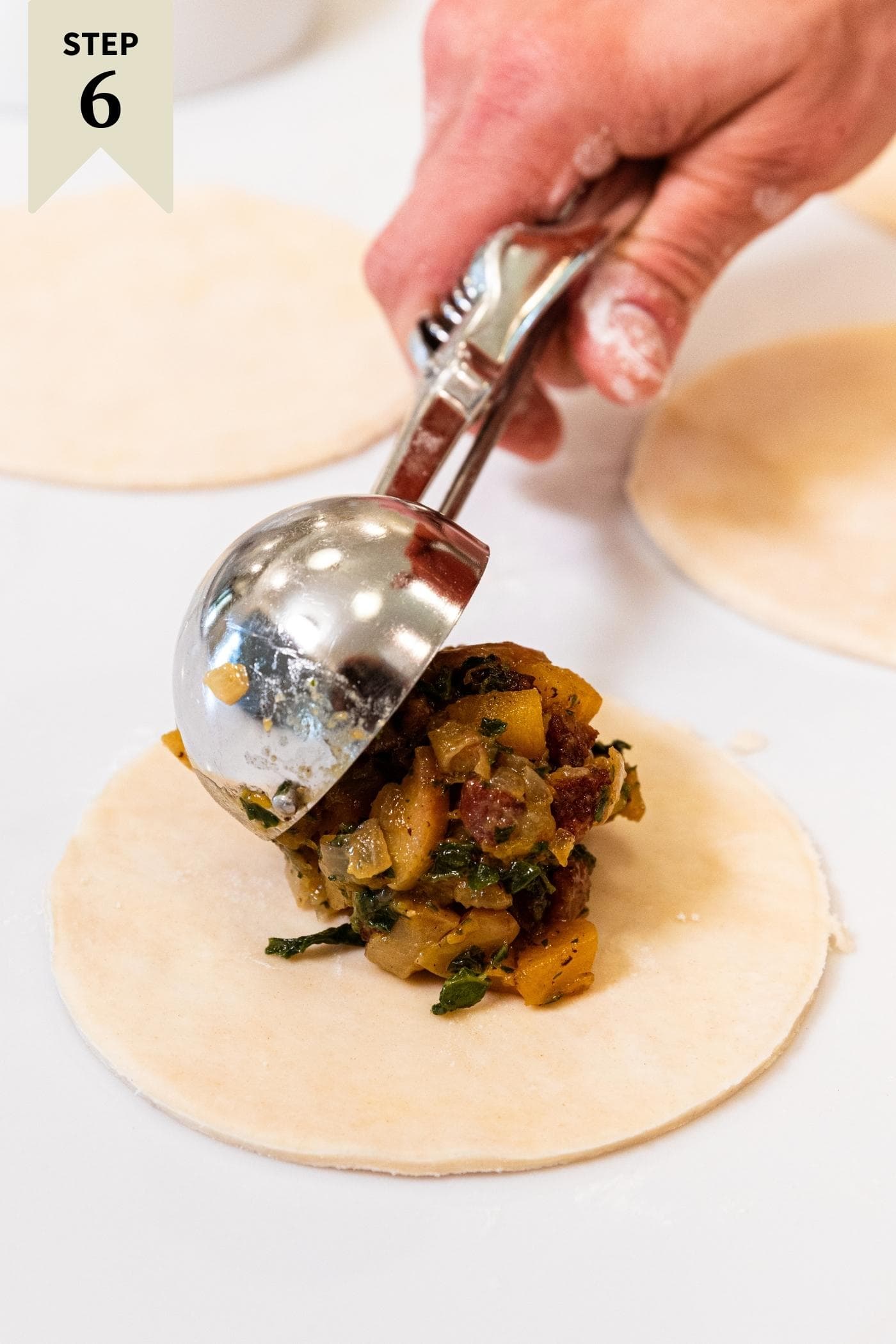 Hand using metal cookie scoop to fill up a round of dough with butternut squash and sausage mixture on white countertop.