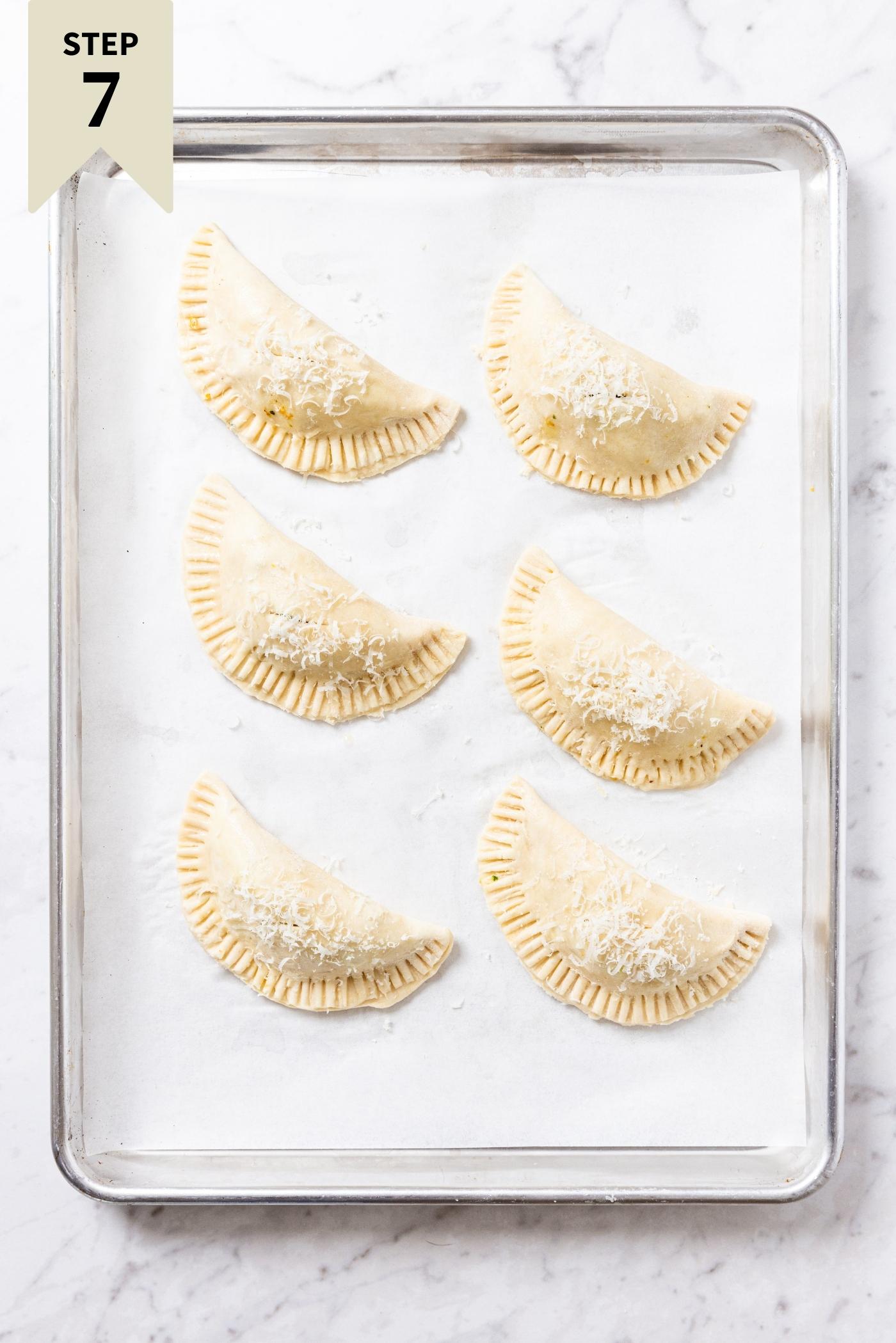 Unbaked half moon shaped hand pies sitting on a white parchment-lined baking sheet sitting on marble countertop before going into the oven to bake.