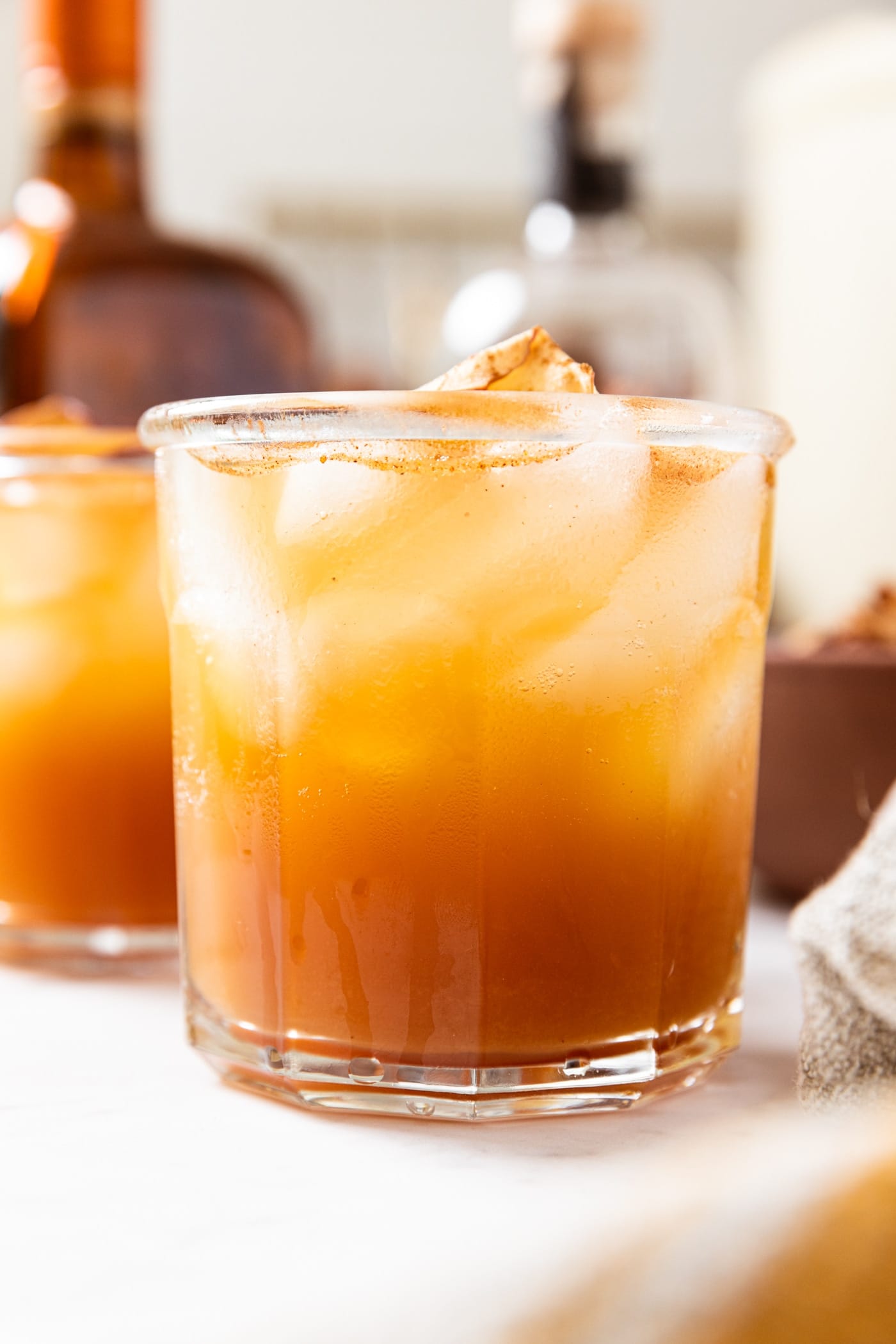 Large glass filled with brown colored apple cider cocktail sitting on marble surface with glass containers filled with more alcohol in background.