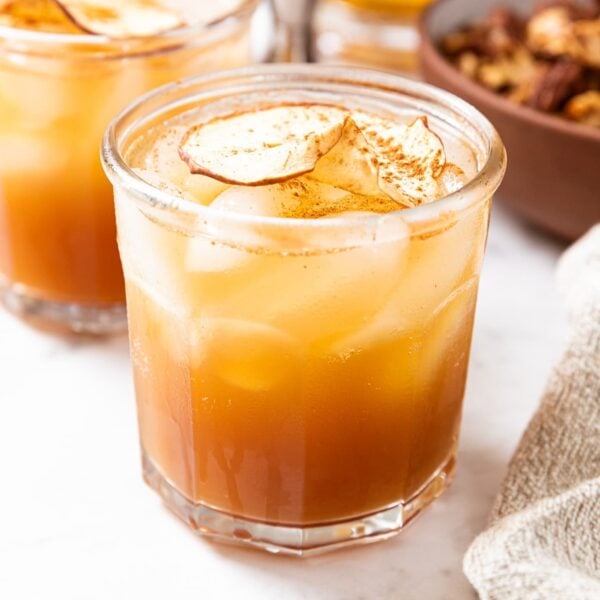 Chunky glass filled with brown colored apple cider sitting on marble surface with bowl full of spiced nuts in background as well as second glass full of same liquid.