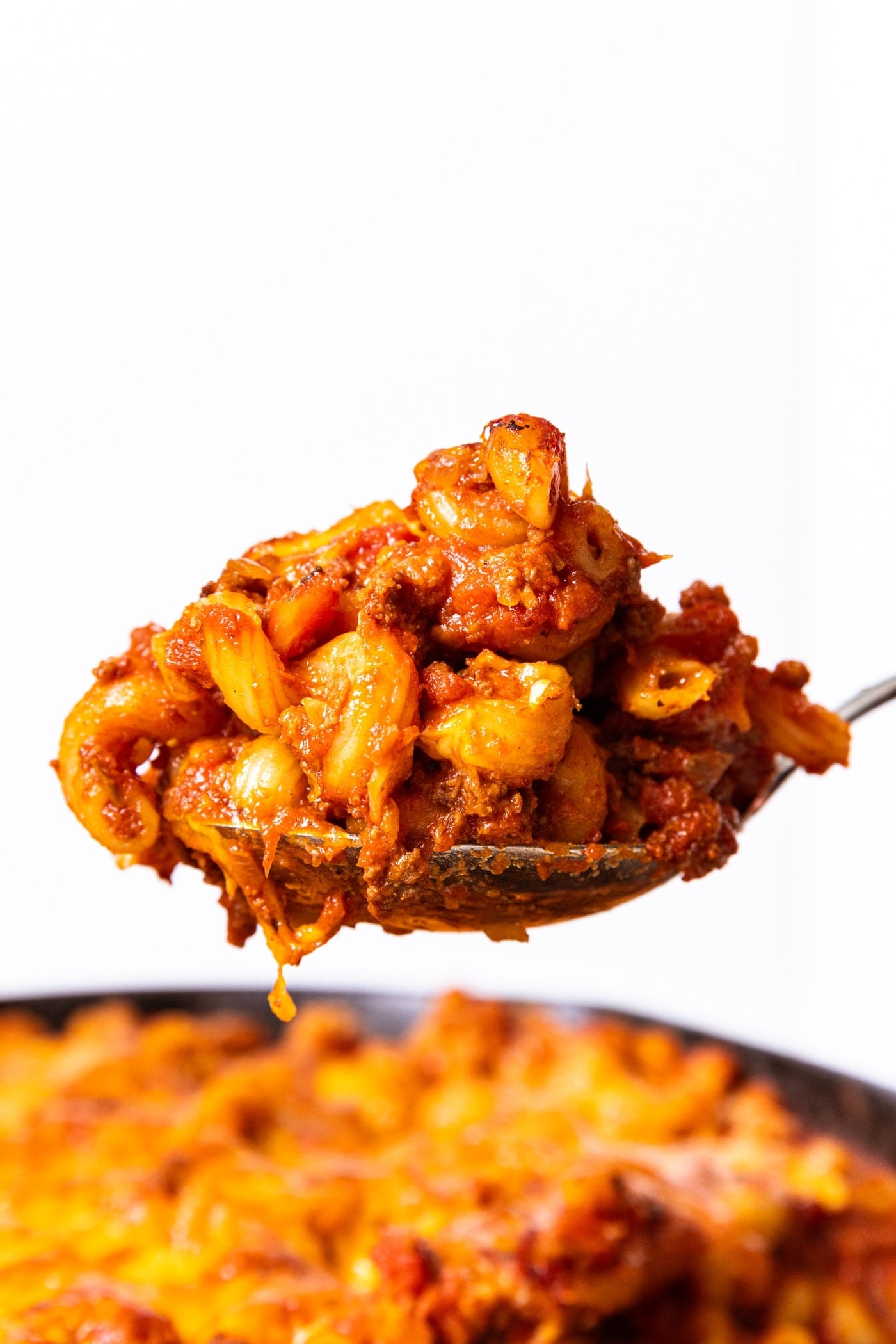 Spoon holding spoonful of homemade goulash with rest of goulash sitting in black cast iron skillet below with white background behind.