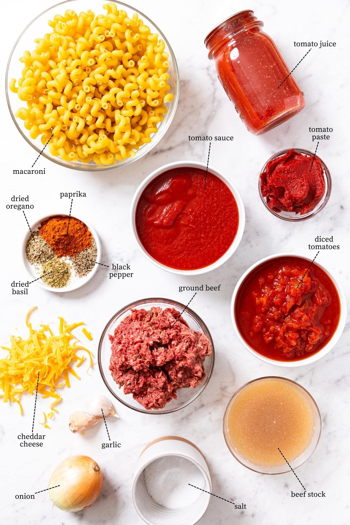 Marble surface with all of the ingredients needed to make homemade goulash including tomato sauces, macaroni, cheese, raw beef, spices, and more.