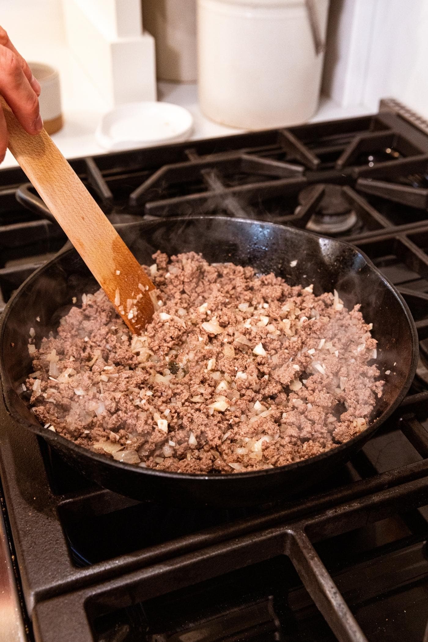 Hand holding wooden spatula mixing together ground beef and onion mixture on stovetop in black cast iron skillet after the meat was browned.