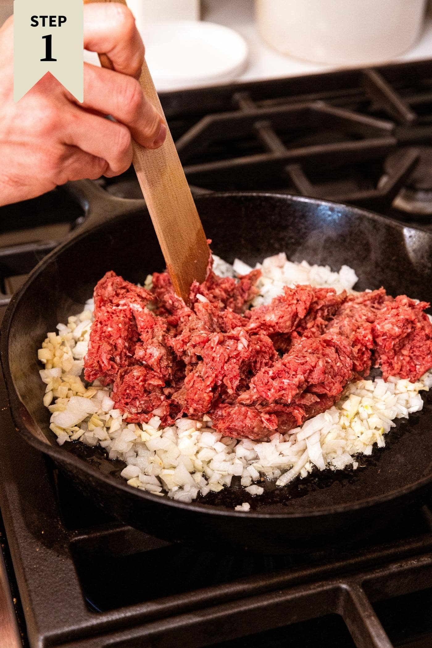 Raw ground beef sitting on top of onions in a black cast iron skillet on stovetop with hand holding wooden spatula mixing together the ingredients.