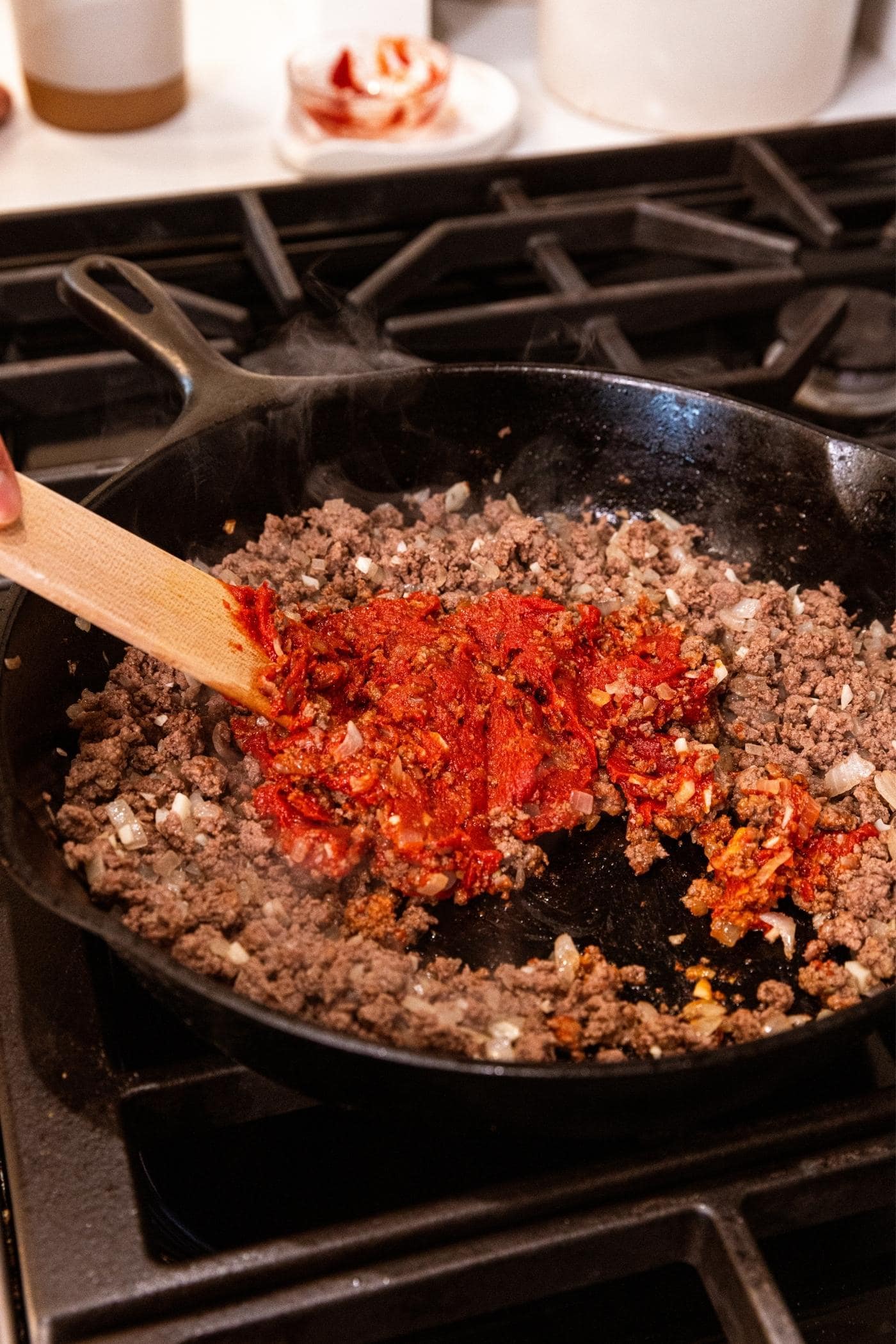 Hand using wooden spatula to mix together tomato paste and browned ground beef in black cast iron skillet.