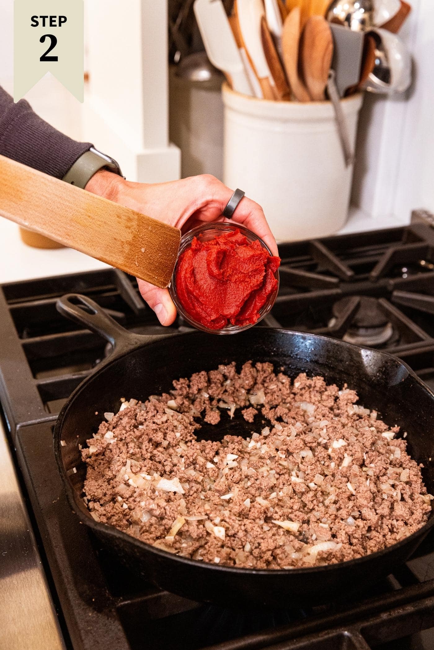 Hand holding small glass bowl with red colored tomato paste with wooden spatula about to scoop out paste into browned ground beef in skillet below.