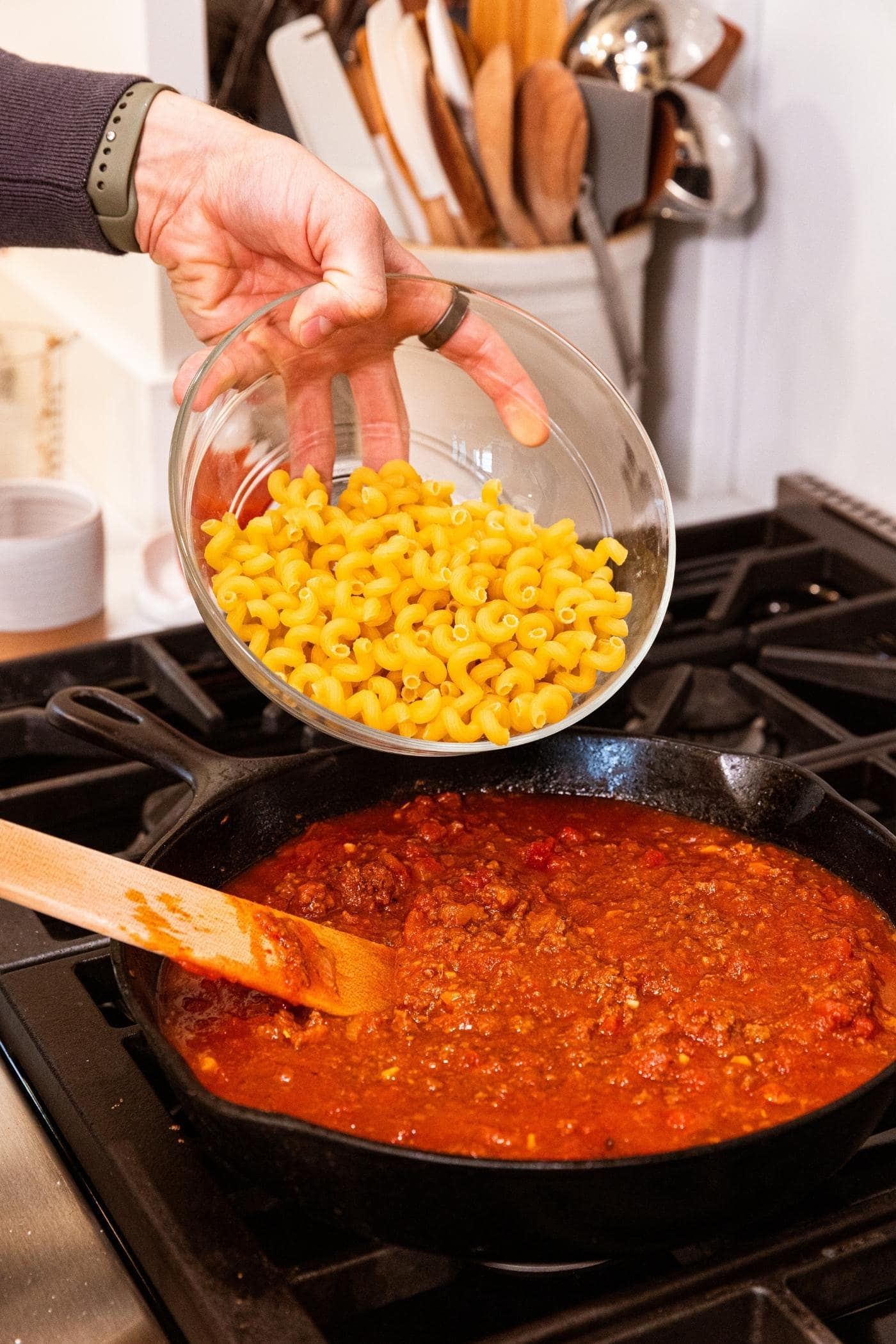 Hand holding glass bowl filled with macaroni about ready to pour into a black cast iron skillet filled with red colored ground beef mixture on stovetop.