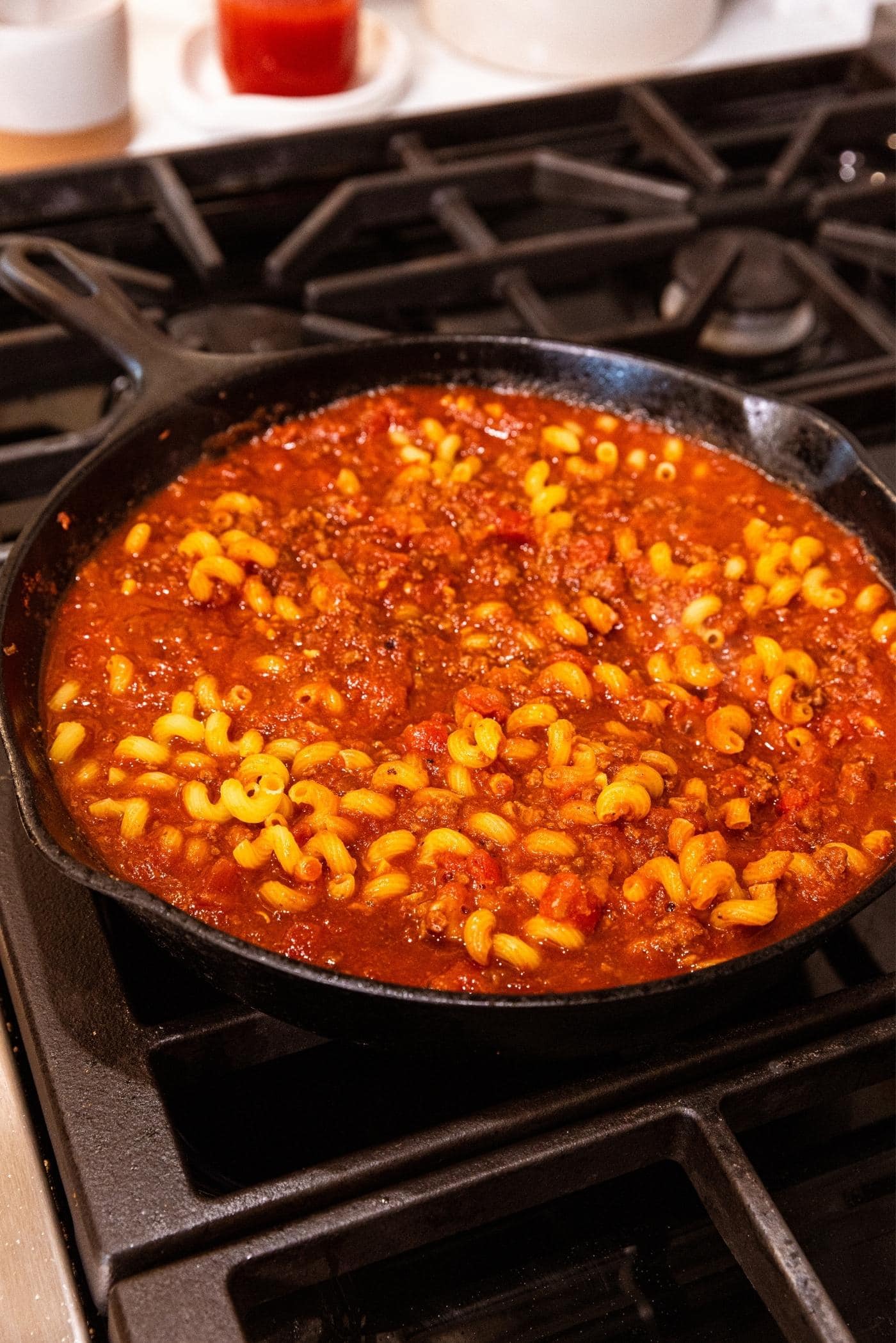Yellow colored macaroni noodles sitting in red colored liquid with ground beef on stovetop in black cast iron skillet.