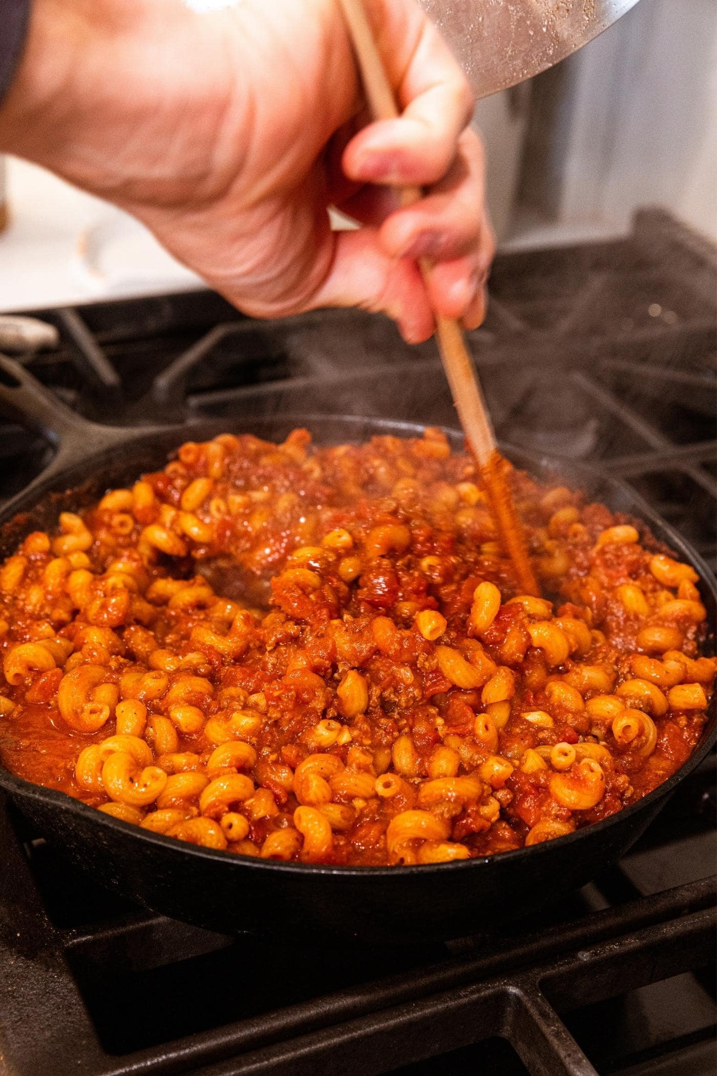 Hand using wooden spatula to mix together red colored ground beef mixture with macaroni noodles in cast iron skillet on stovetop.