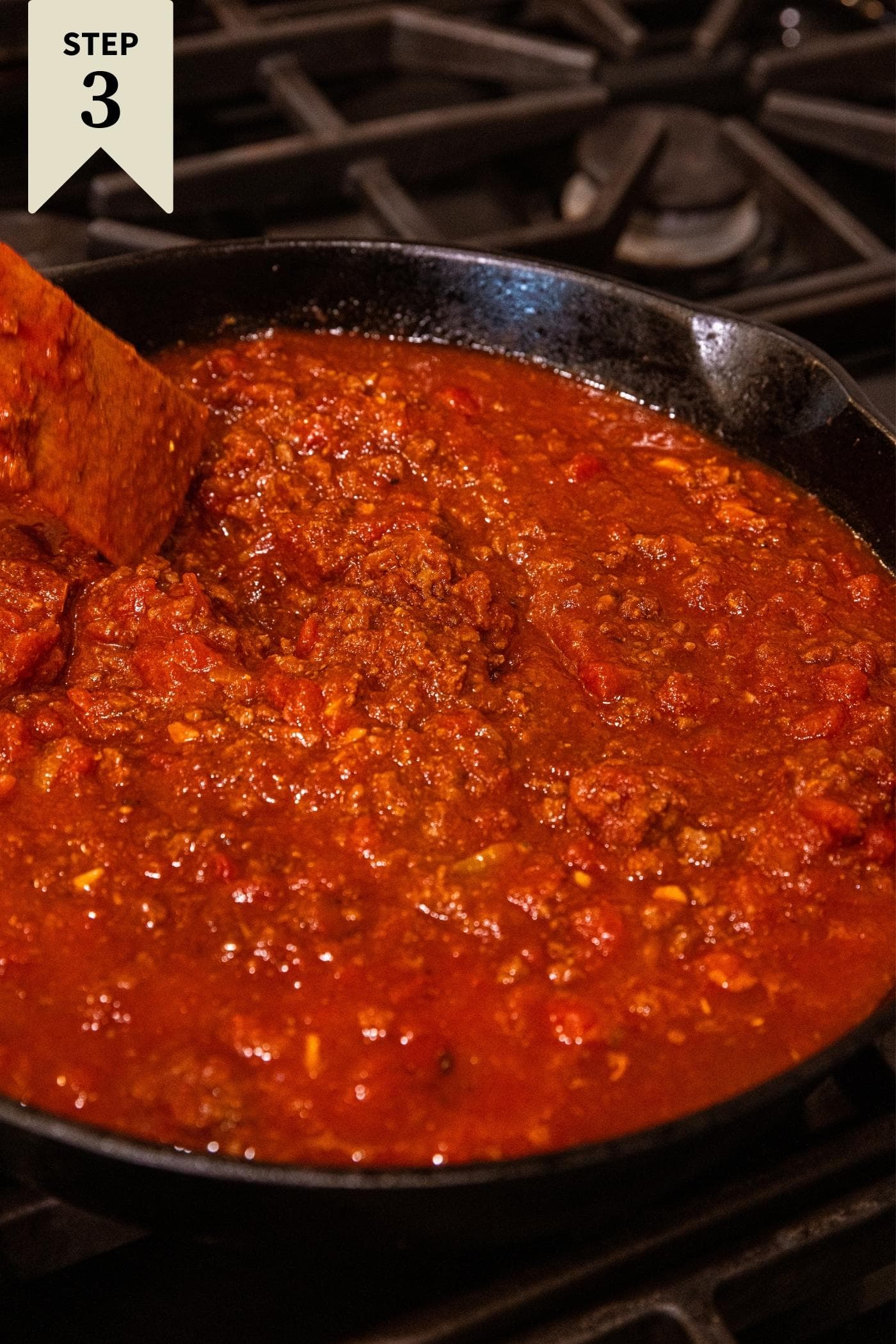 Red colored liquid ground beef mixture sitting in black cast iron skillet on stovetop after simmering for some time with wooden spatula resting in skillet.
