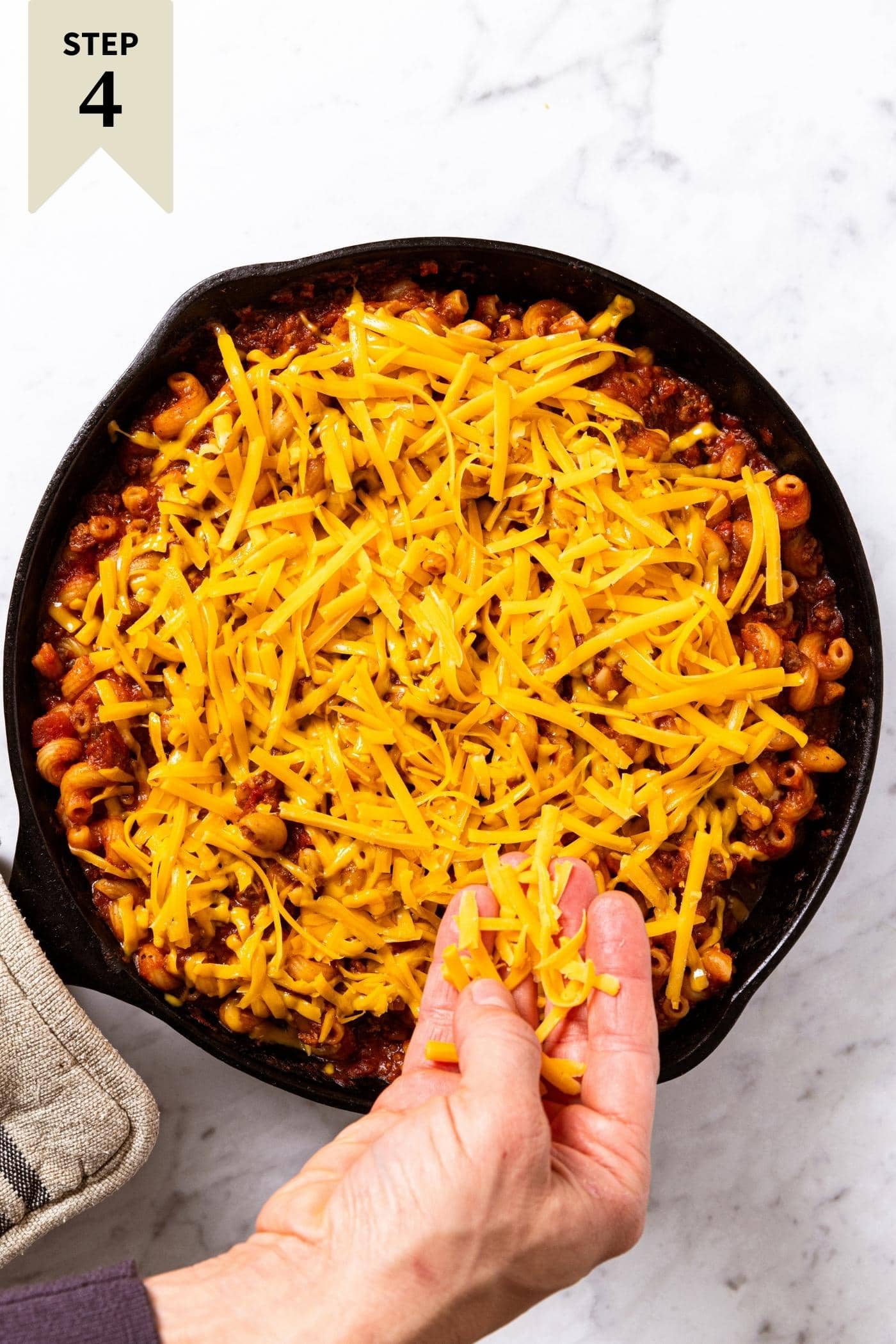 Hand sprinkling unmelted yellow cheddar cheese all over the top of ground beef mixture sitting in black cast iron skillet before being baked in oven.