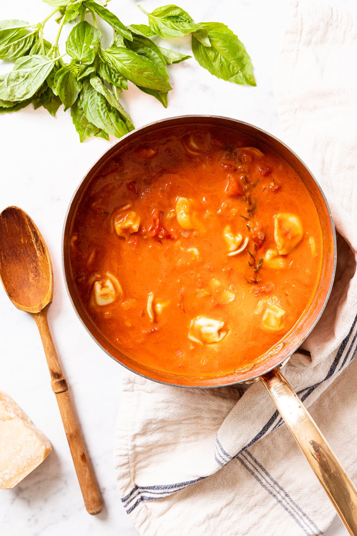 Copper pot sitting on marble surface filled with tomato tortellini soup with wooden spoon sitting beside pot along with fresh basil and Parmesan cheese.