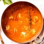 Marble surface with copper pot filled with tomato tortellini soup with wooden spoon beside and cooked sprig of rosemary on top of soup surface.