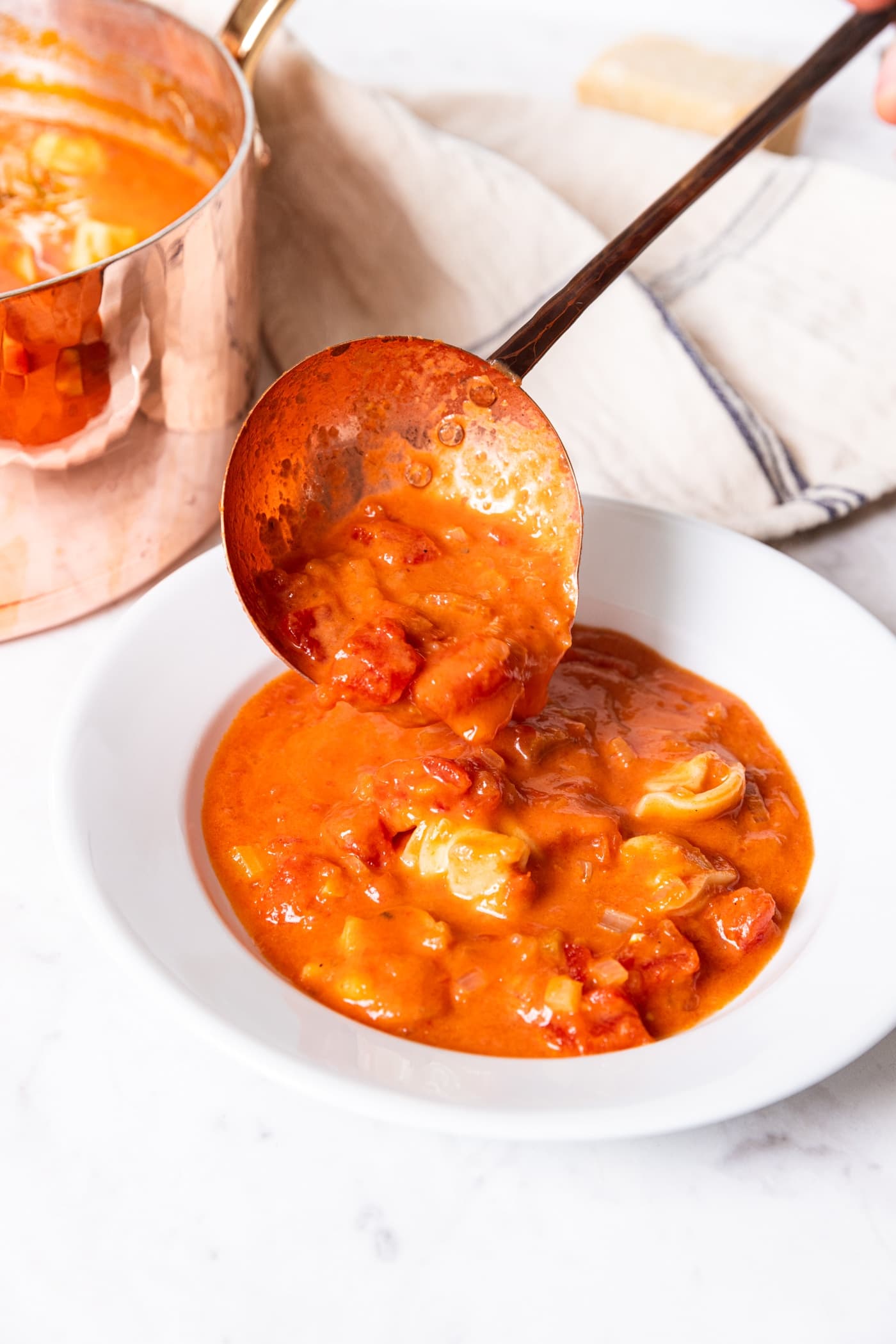 Ladle serving up tomato tortellini soup in a white bowl on a white marble surface.