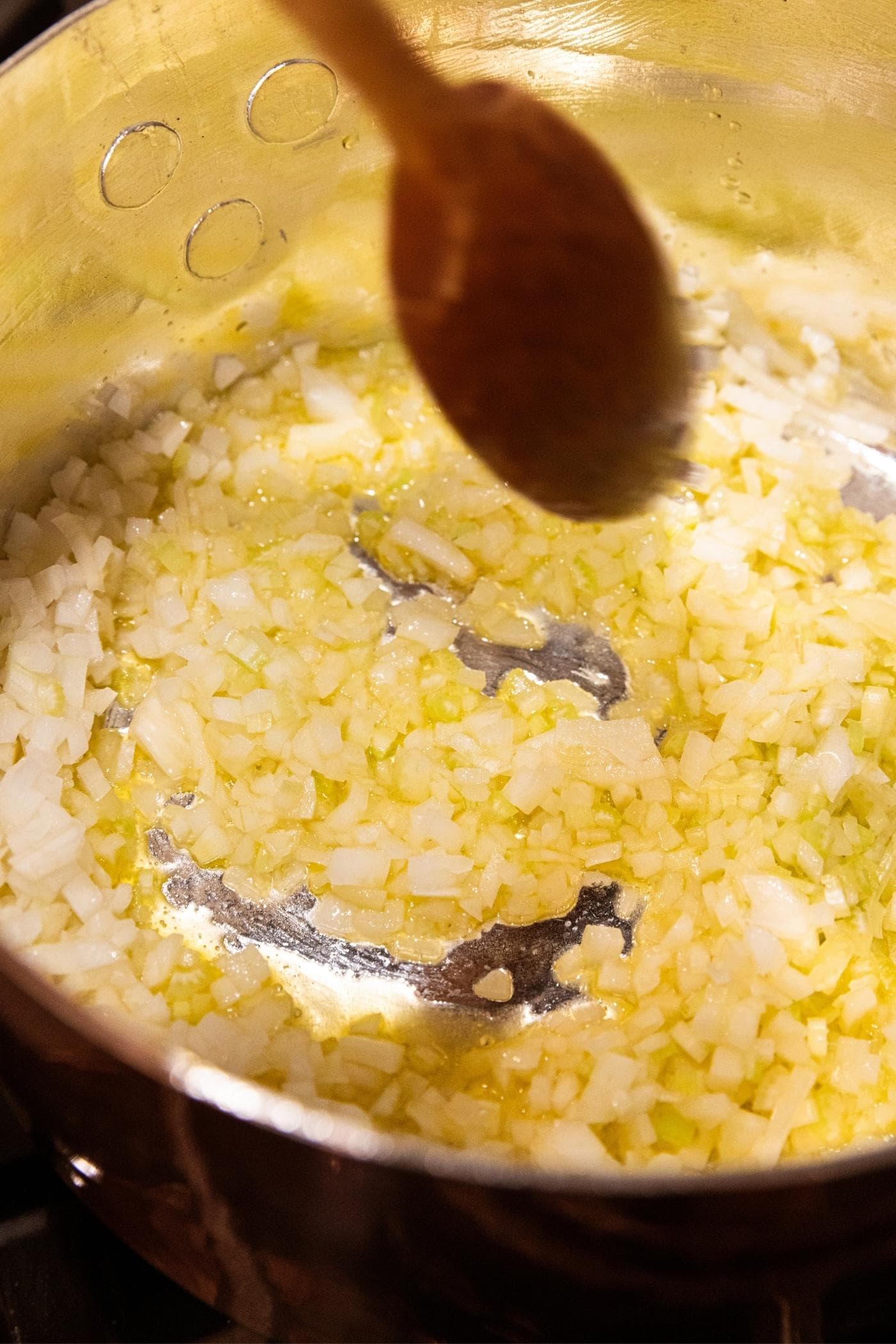 Metal pot sitting on the stovetop with onion and celery mixture sautéing with wooden spoon mixing everything together.