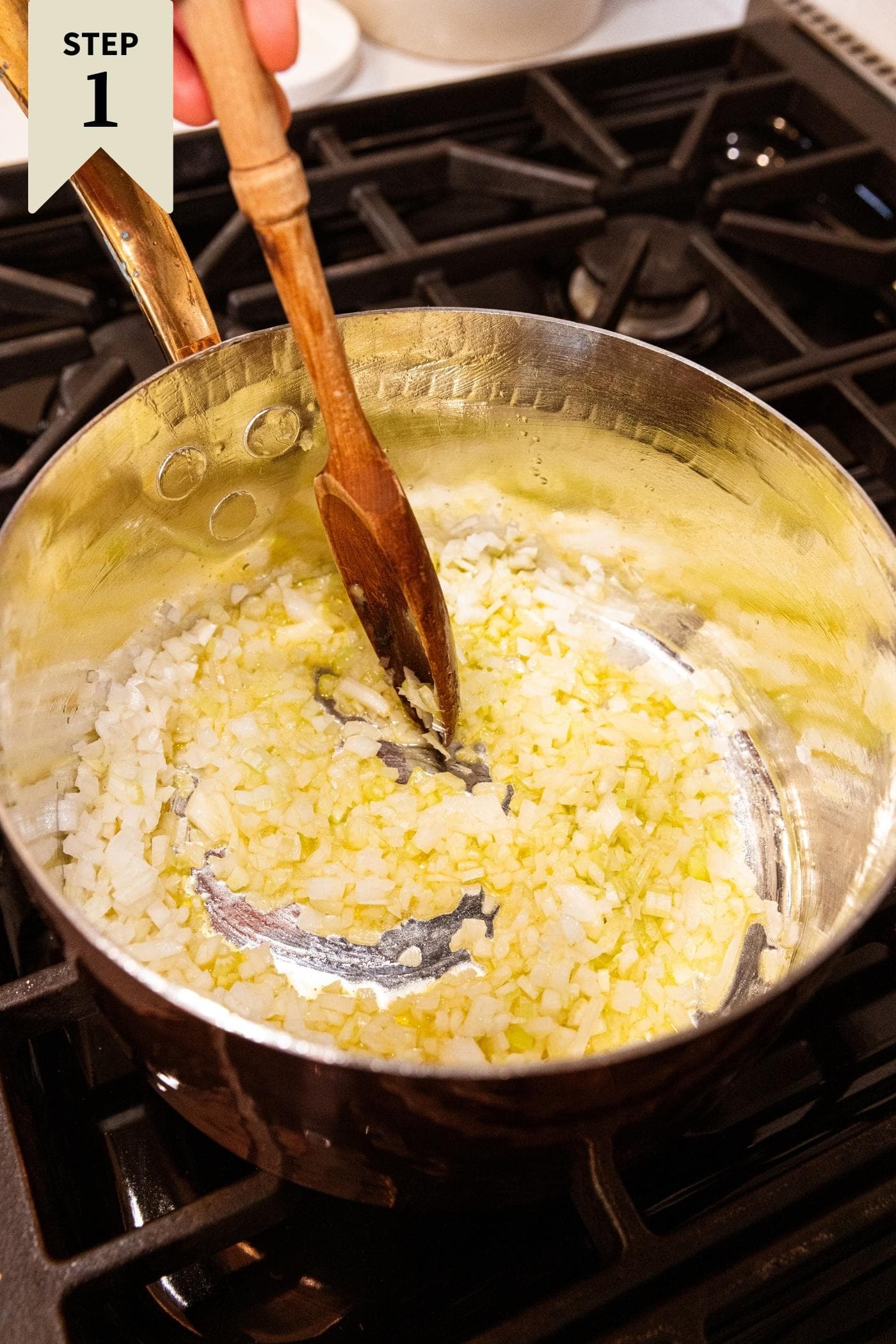 Wooden spoon mixing together onion and celery in copper pot on stovetop.
