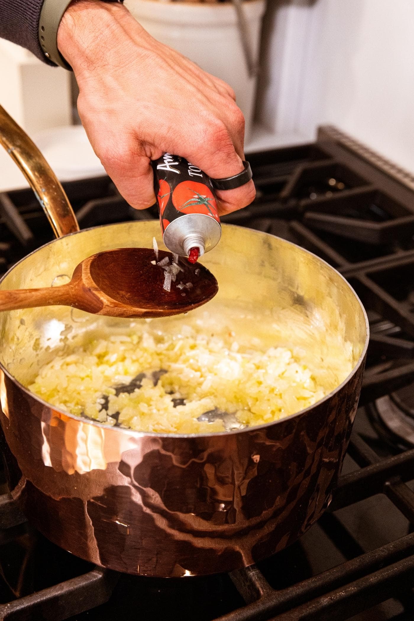 Hand holding wooden spoon with tomato paste being squeezed from bottle onto spoon with celery and onion mixture in kettle below.