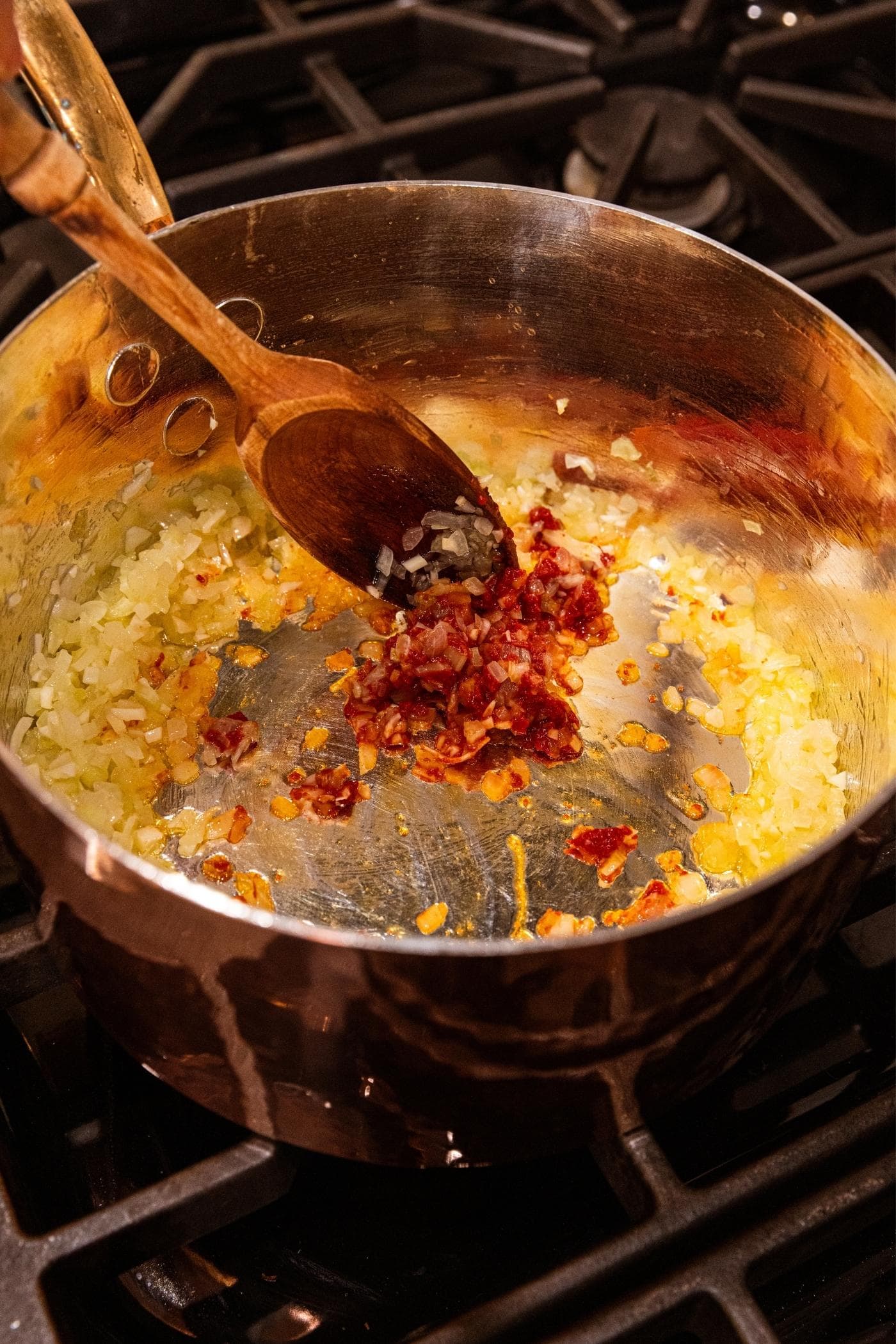 Wooden spoon stirring together tomato paste into celery and onion mixture in a copper pot on stovetop.