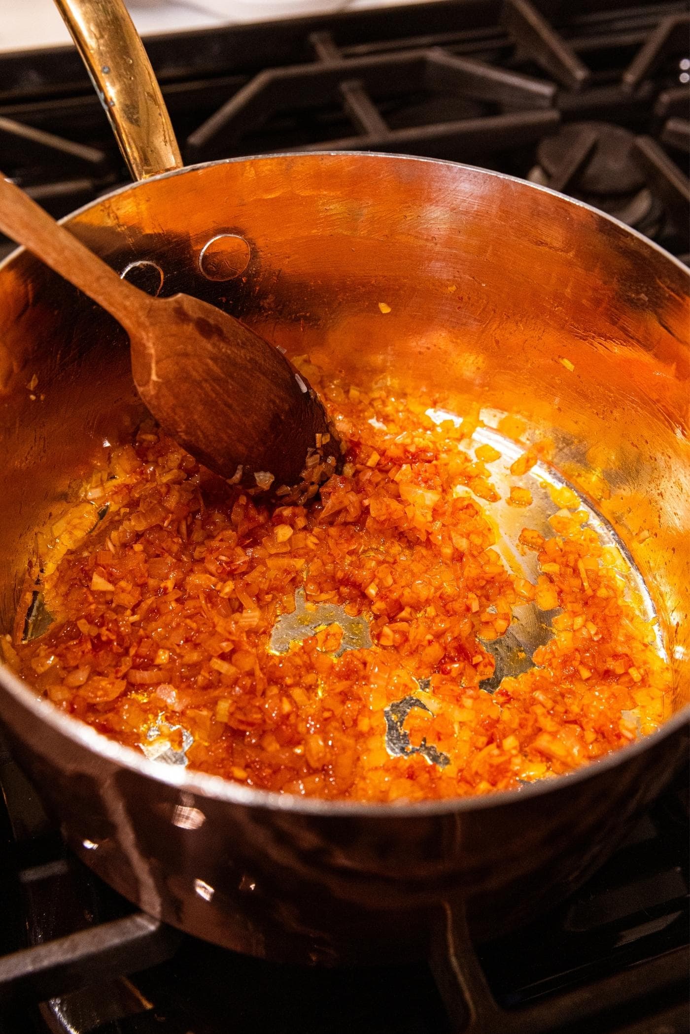 Tomato paste worked together with onion and celery mixture in copper pot on stovetop with wooden spoon mixing everything together.