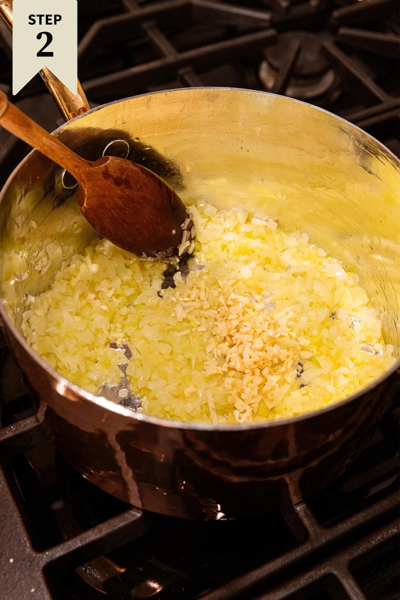 Chopped garlic about to be mixed into a celery and onion mixture in copper pot sitting on stovetop with wooden spoon about to mix.