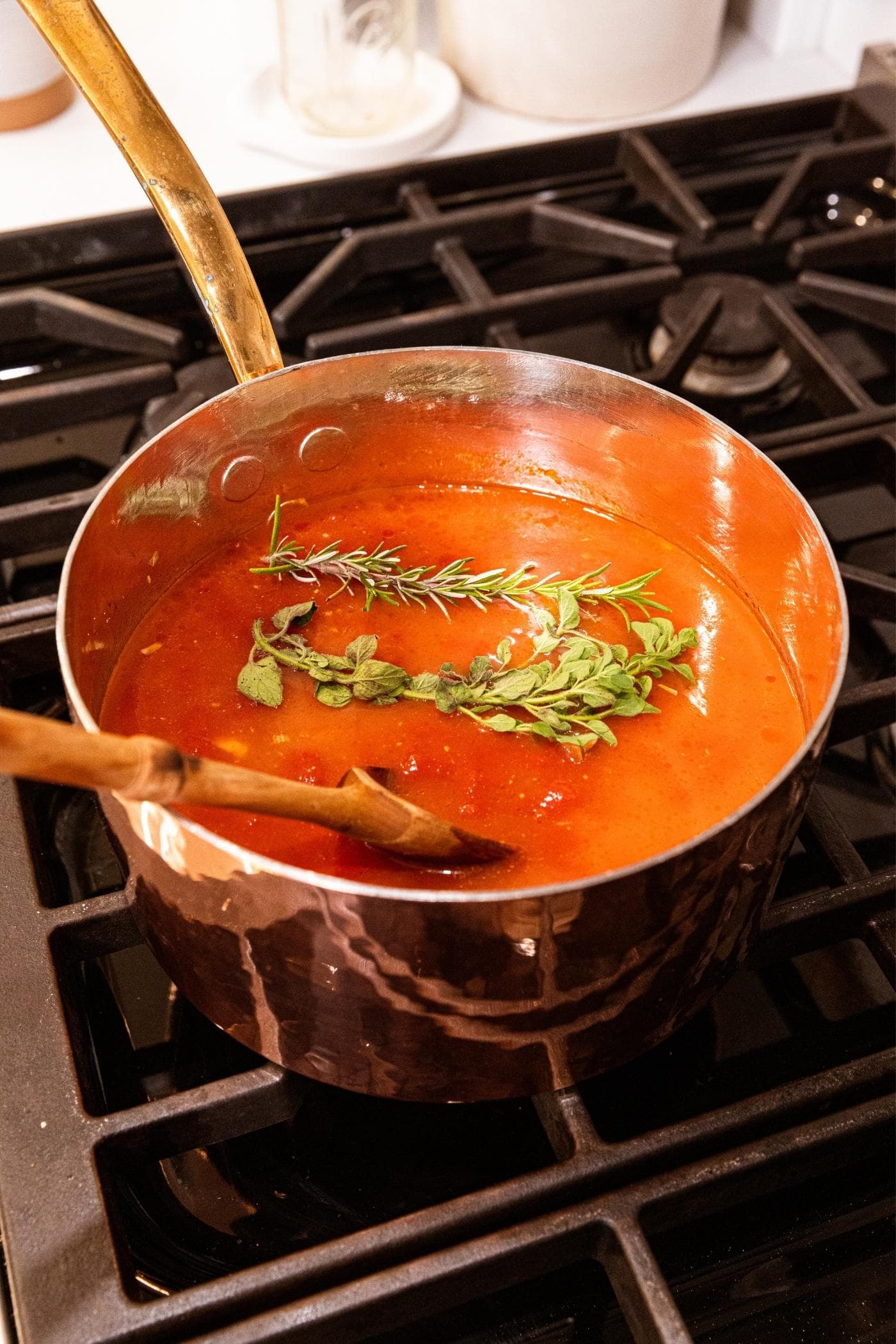 Copper pot sitting on stovetop with tomato soup in kettle with rosemary and thyme sitting on top before sinking into soup.