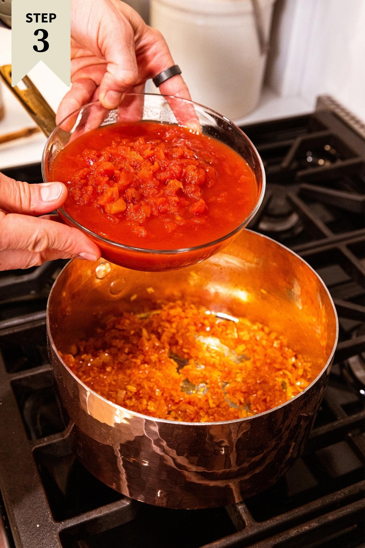 Two hands holding glass bowl filled with diced tomatoes before pouring into a copper pot filled with tomato paste and onion and celery mixture.