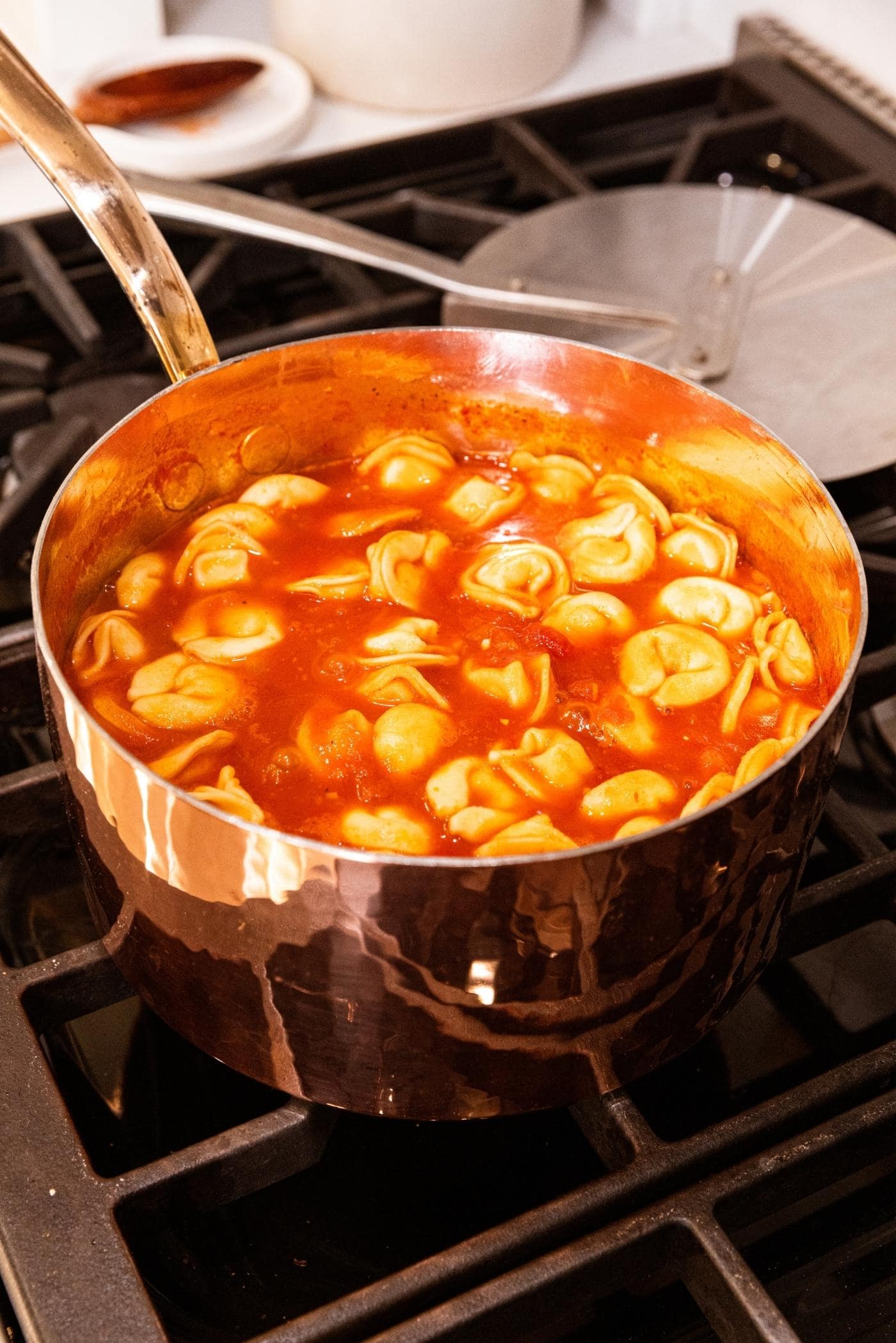 Partially cooked tomato tortellini sitting in copper pot amongst tomato soup with metal lid in background.
