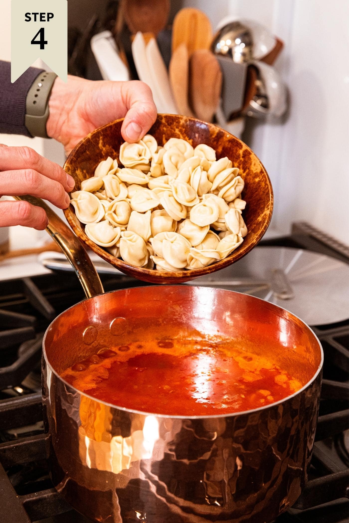 Hand holding brown bowl filled with uncooked tortellini before pouring into red tomato soup mixture on stovetop.