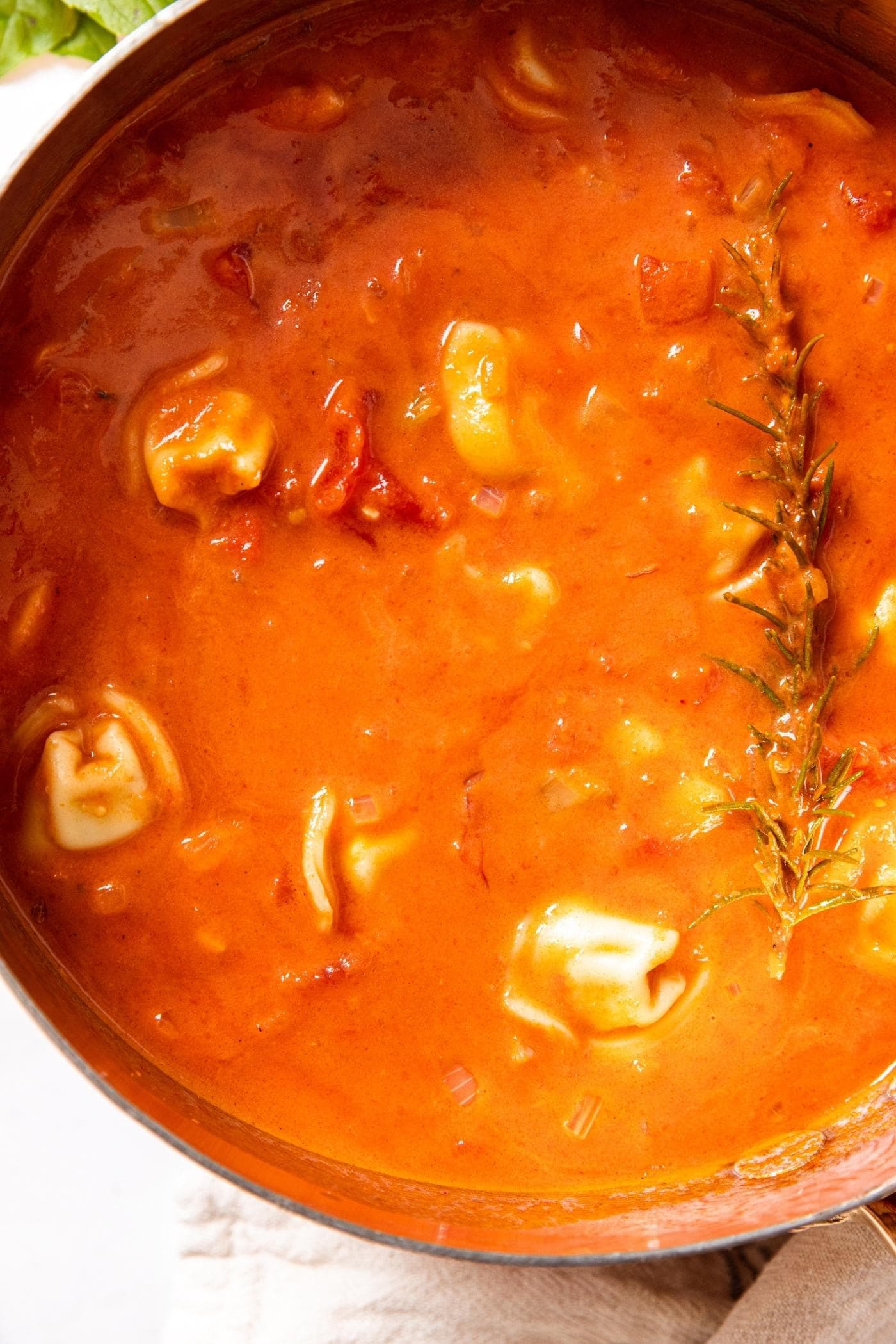 Copper pot filled with tomato tortellini soup with cooked sprig of rosemary resting on the top of the soup all on white marble surface.