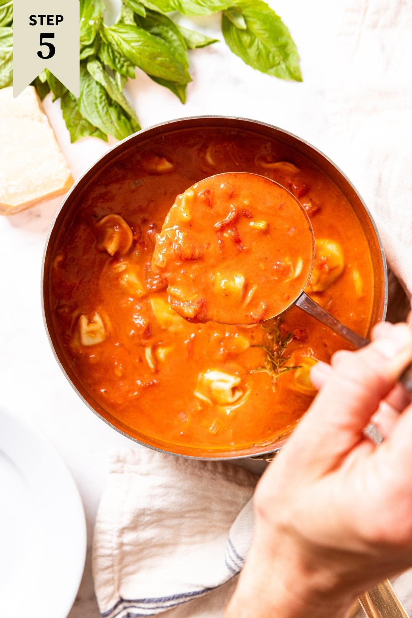 Marble surface with pot of tomato tortellini soup mixed with heavy cream with hand scooping up ladle full of soup about to put in white bowl with fresh basil to the side.