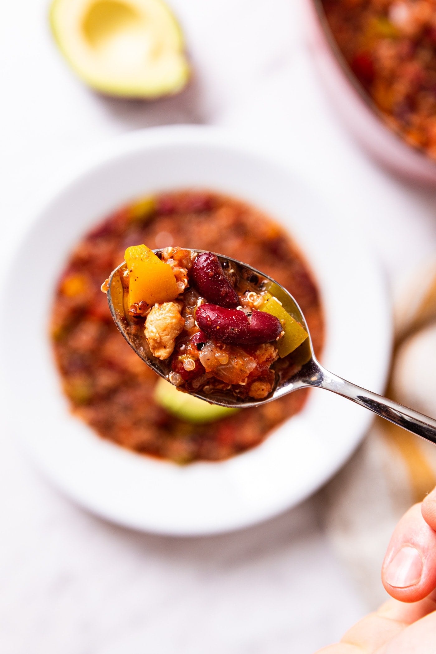 Spoon holding scoop of turkey quinoa chili with white bowl sitting below filled with more chili.
