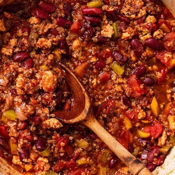 Large red Dutch oven filled with turkey quinoa chili with a wooden spoon resting on the side of the pot with pieces of meat and vegetables inside.