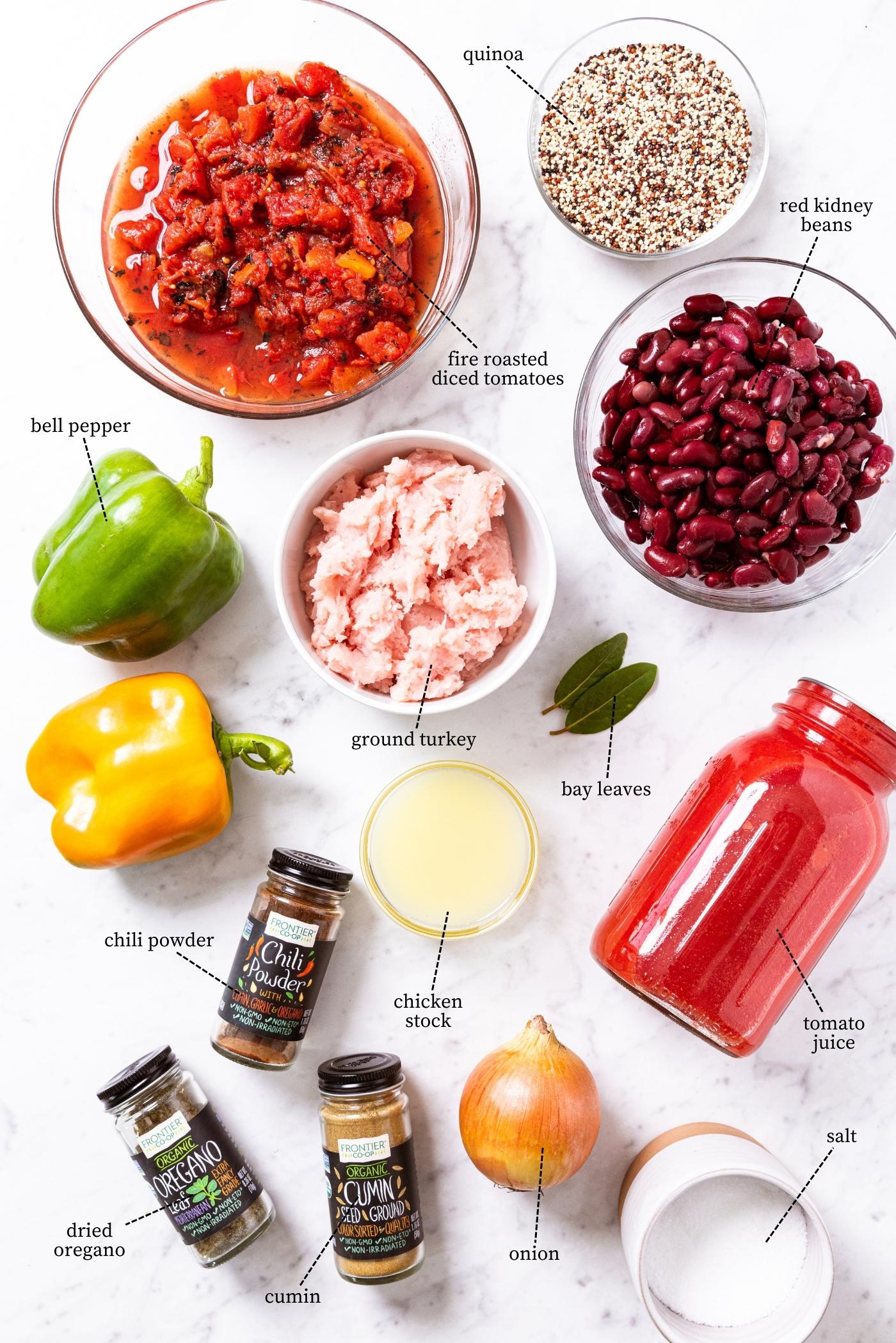 White marble surface filled with all of the ingredients needed to make turkey quinoa chili including tomato juice, bell peppers, ground turkey, beans, diced tomatoes, spices, and more.