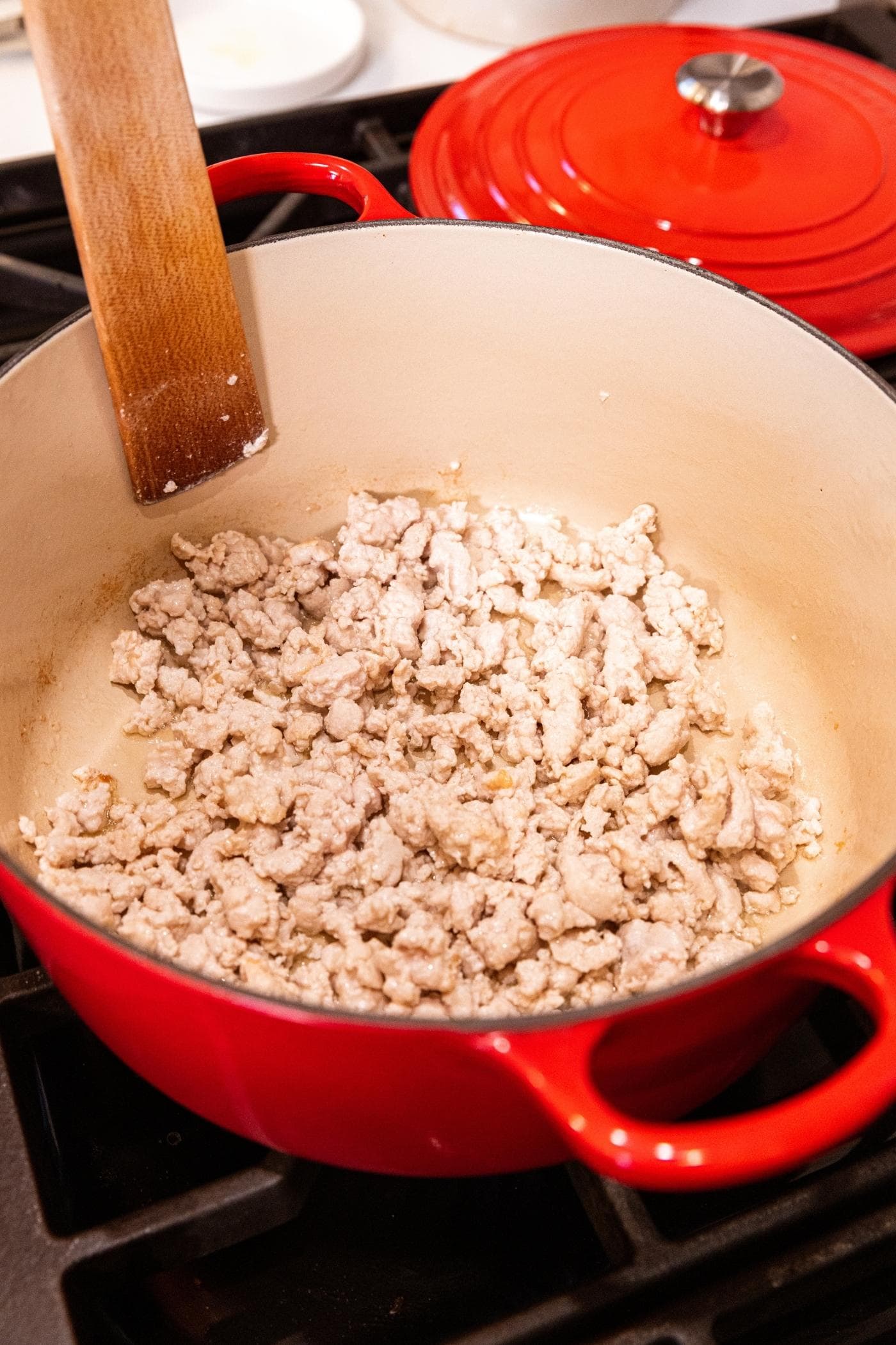 Cooked ground turkey sitting in bottom of red Dutch oven with wooden spatula about to continue pushing around the meat.