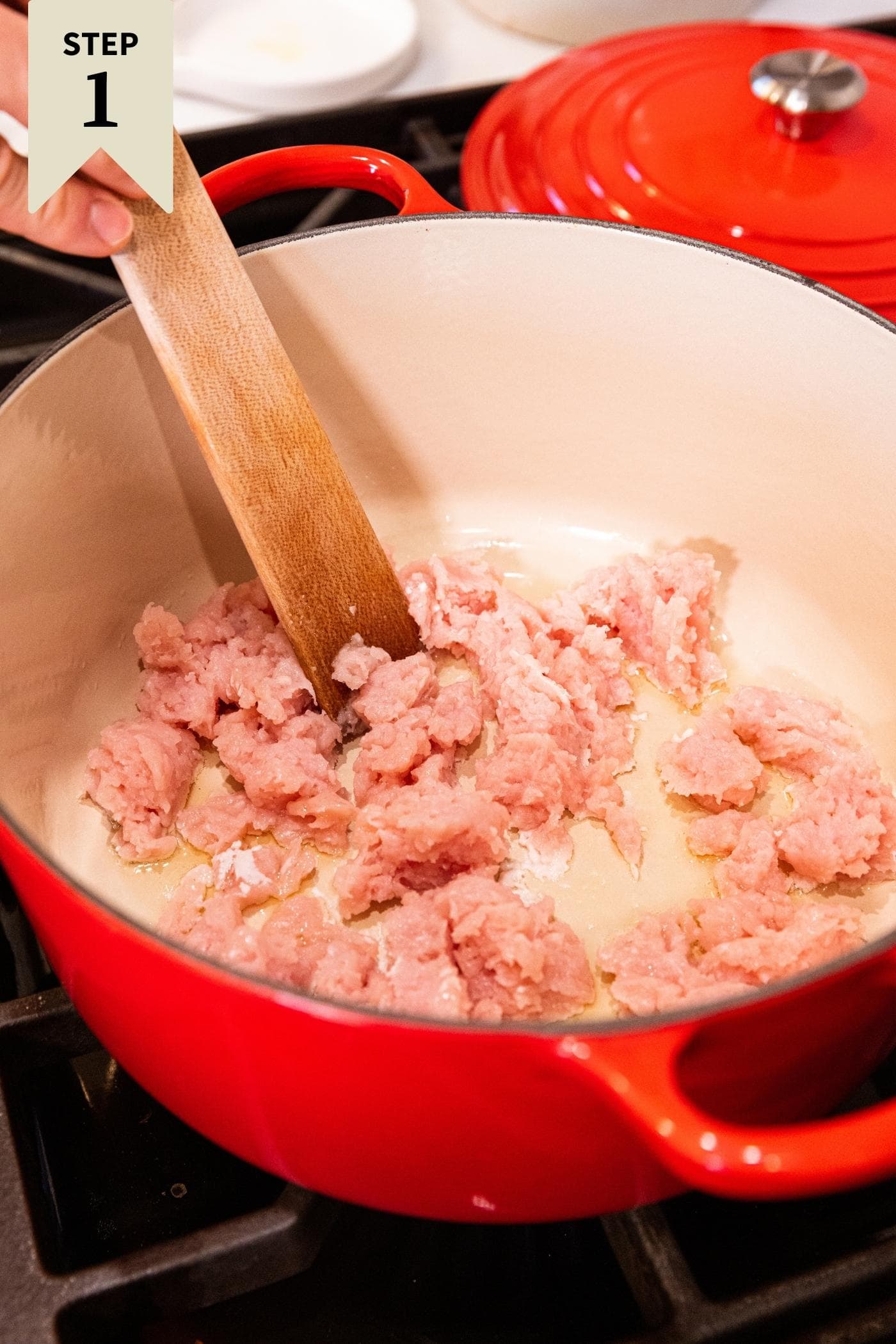 Raw ground turkey meat in red Dutch oven on stovetop with wooden spatula mixing up the meat.