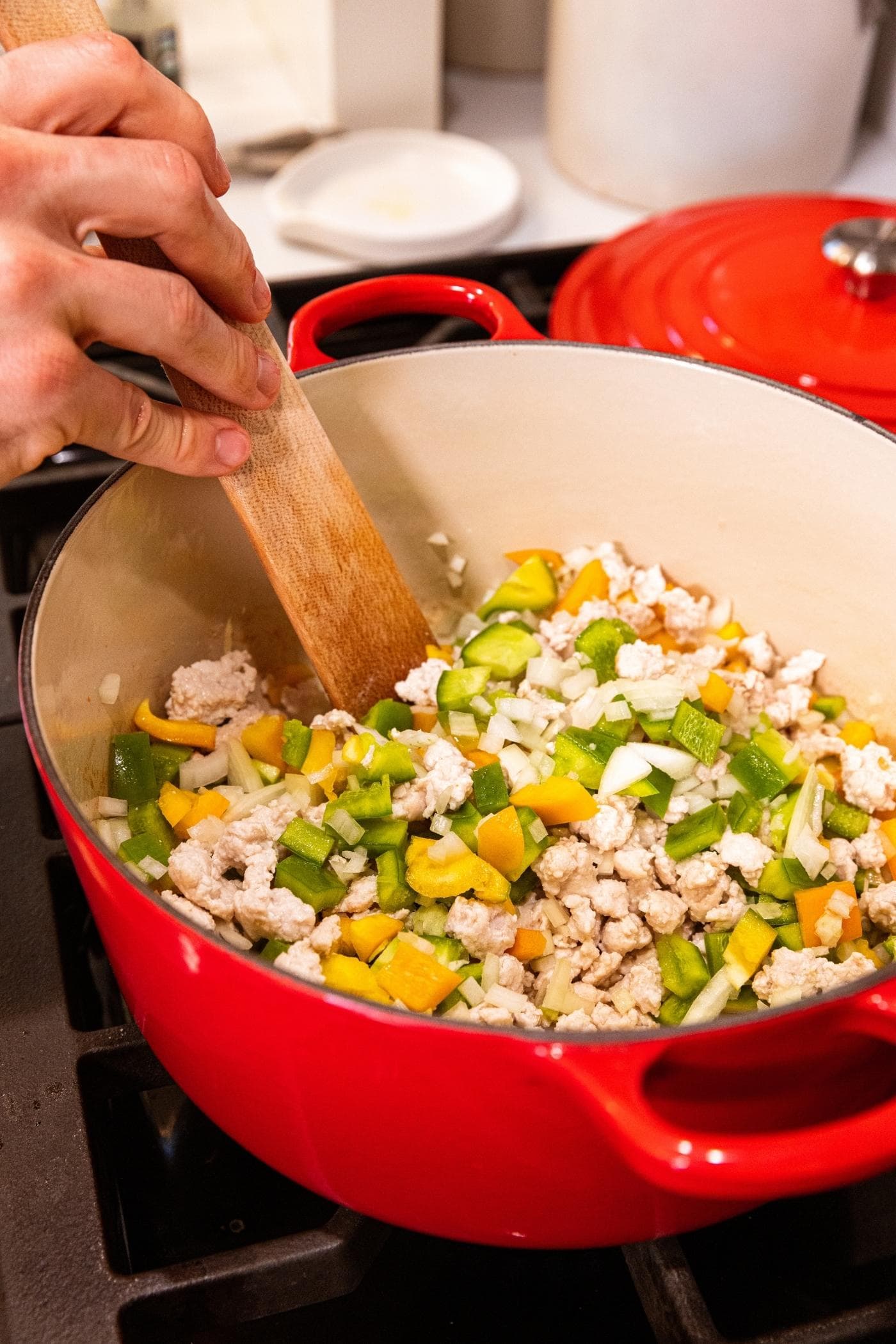 Wooden spatula stirring together diced green and yellow bell pepper and onion with ground turkey in red Dutch oven.